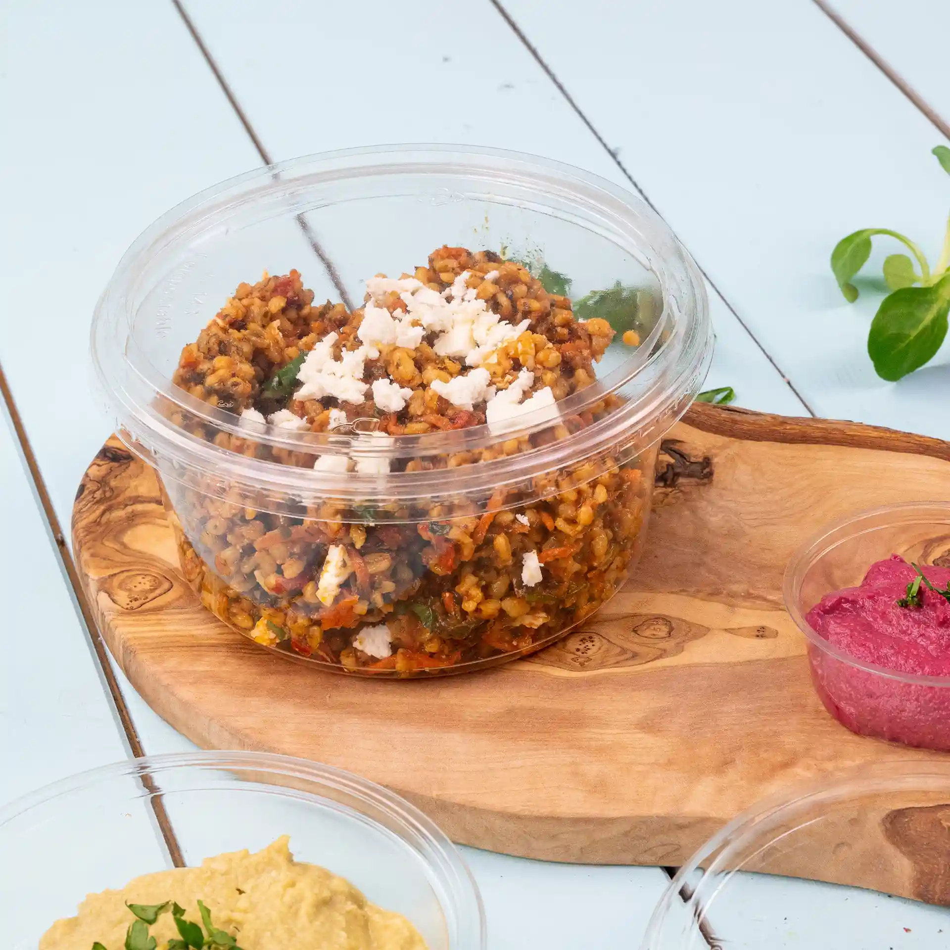 Transparent round deli cup with a lid, containing a grain dish topped with crumbled cheese, placed on a wooden board.