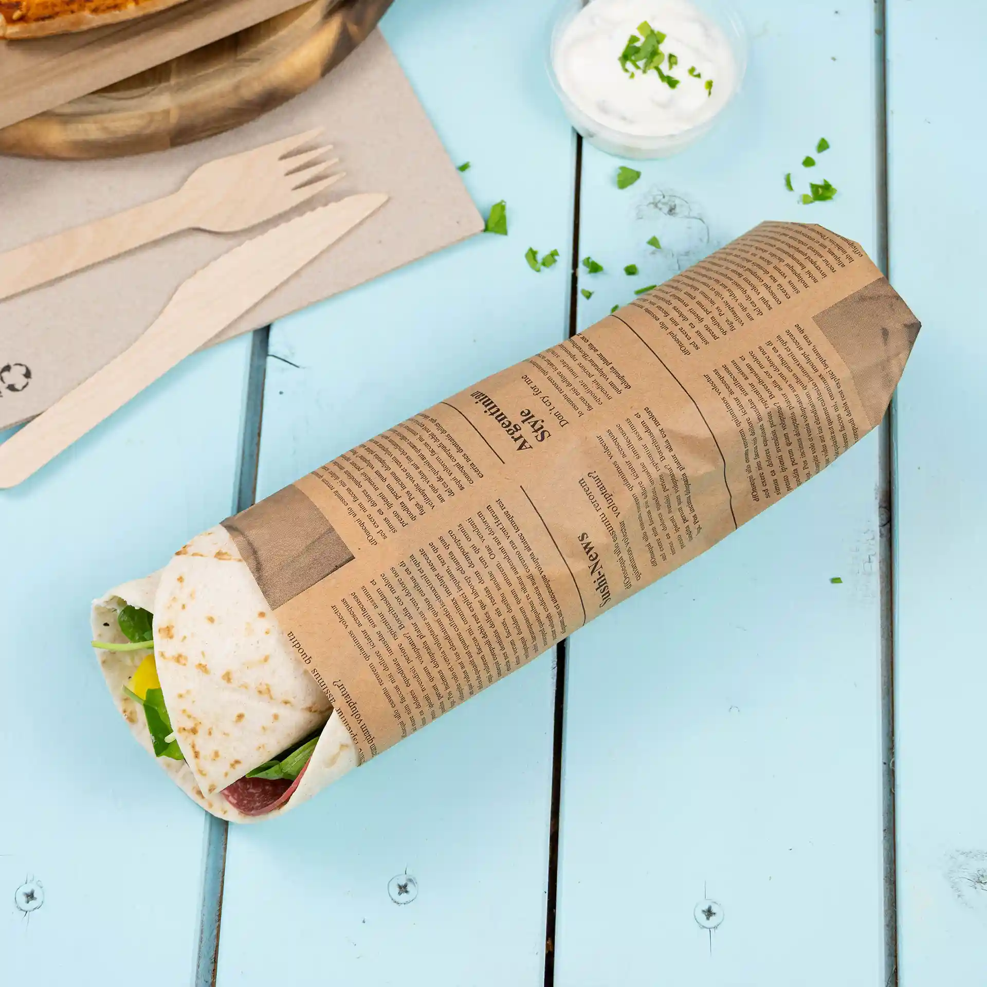 Wrapped food item in printed kraft paper on a light blue wooden surface, with wooden utensils and a small bowl of sauce nearby.