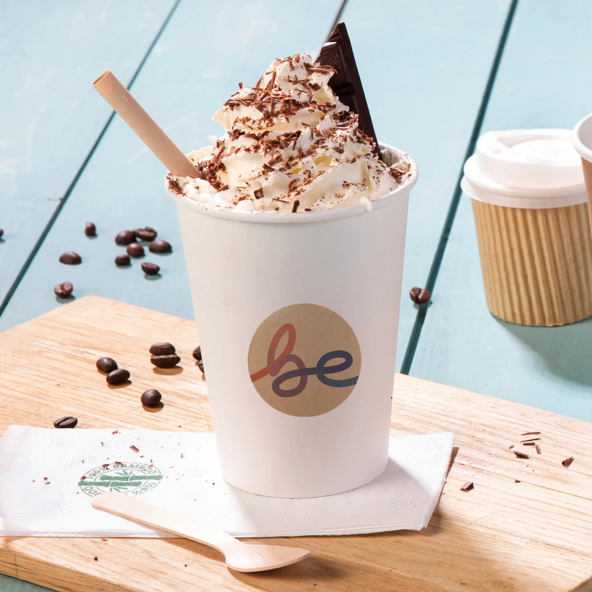 Whipped cream topped drink in a white cup with a "be" logo, surrounded by coffee beans and a wooden spoon.