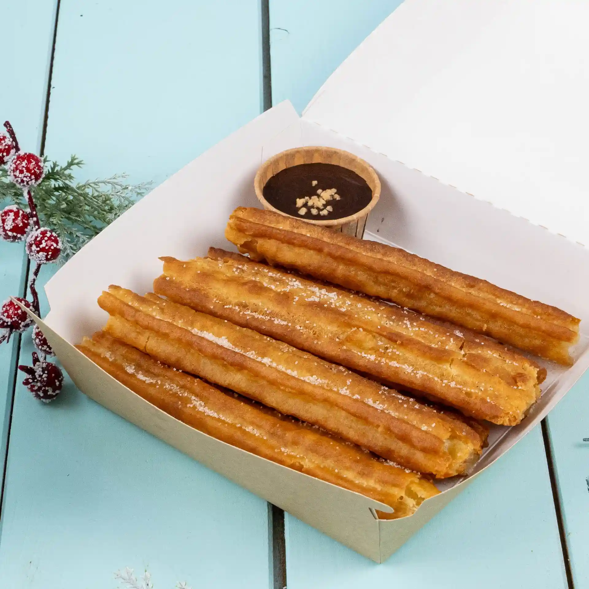 Churros in a paper box with a small brown cup of chocolate sauce on a blue surface.