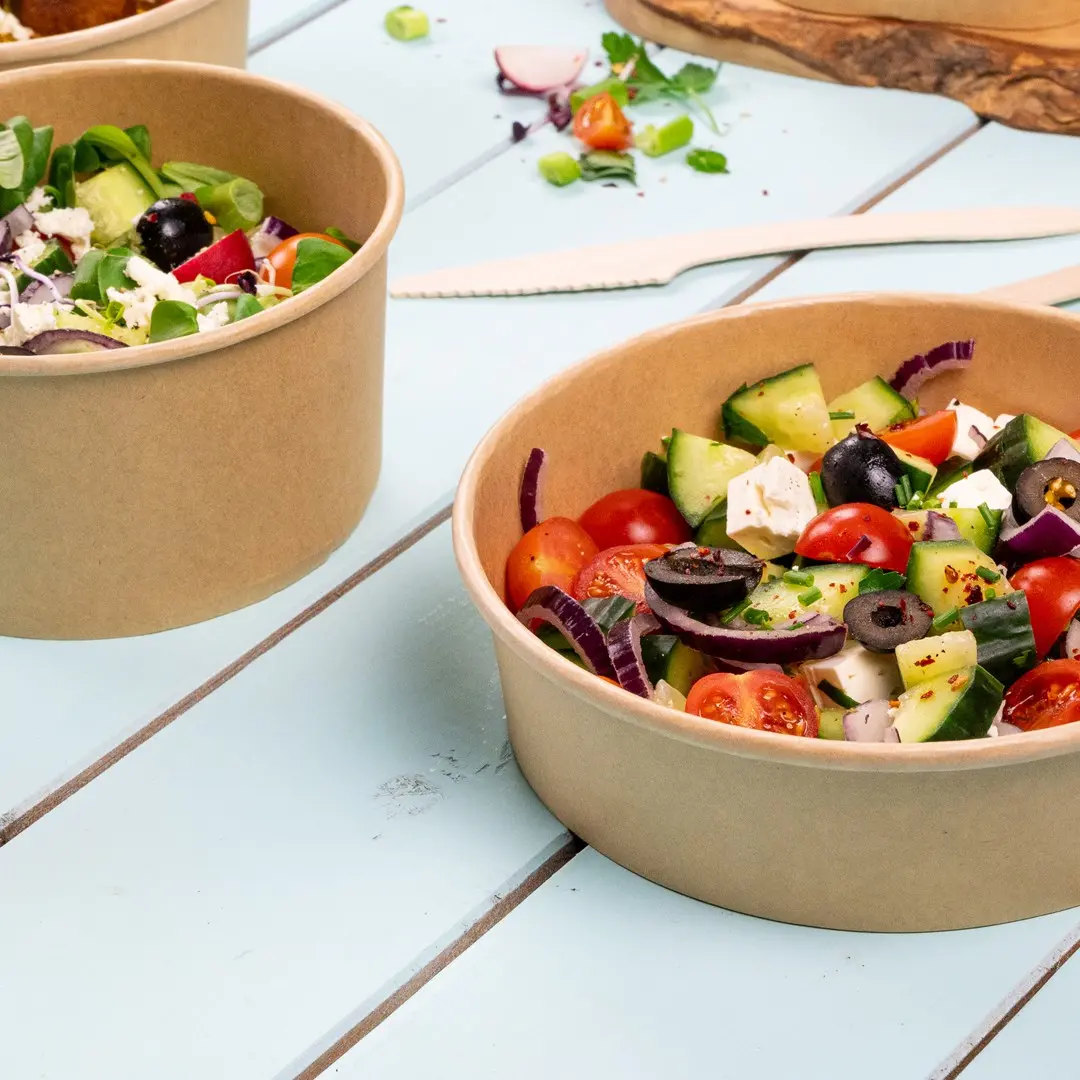 Two brown paper salad bowls filled with mixed vegetables, including tomatoes, cucumbers, olives, and greens, on a blue wooden surface.