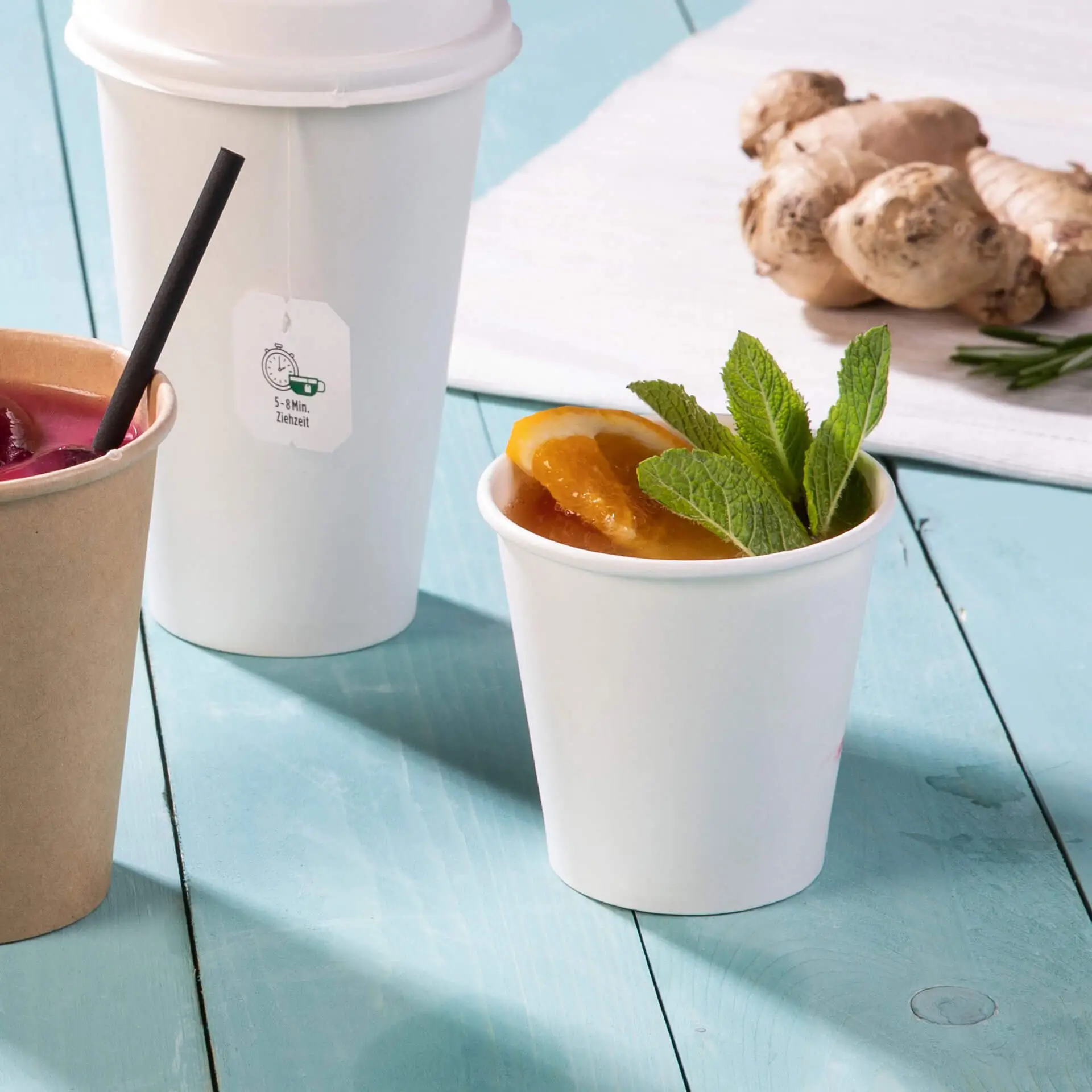 One white paper cup filled with a drink, garnished with mint leaves and orange slices, alongside a brown paper cup and another white cup. Fresh ginger roots are in the background.