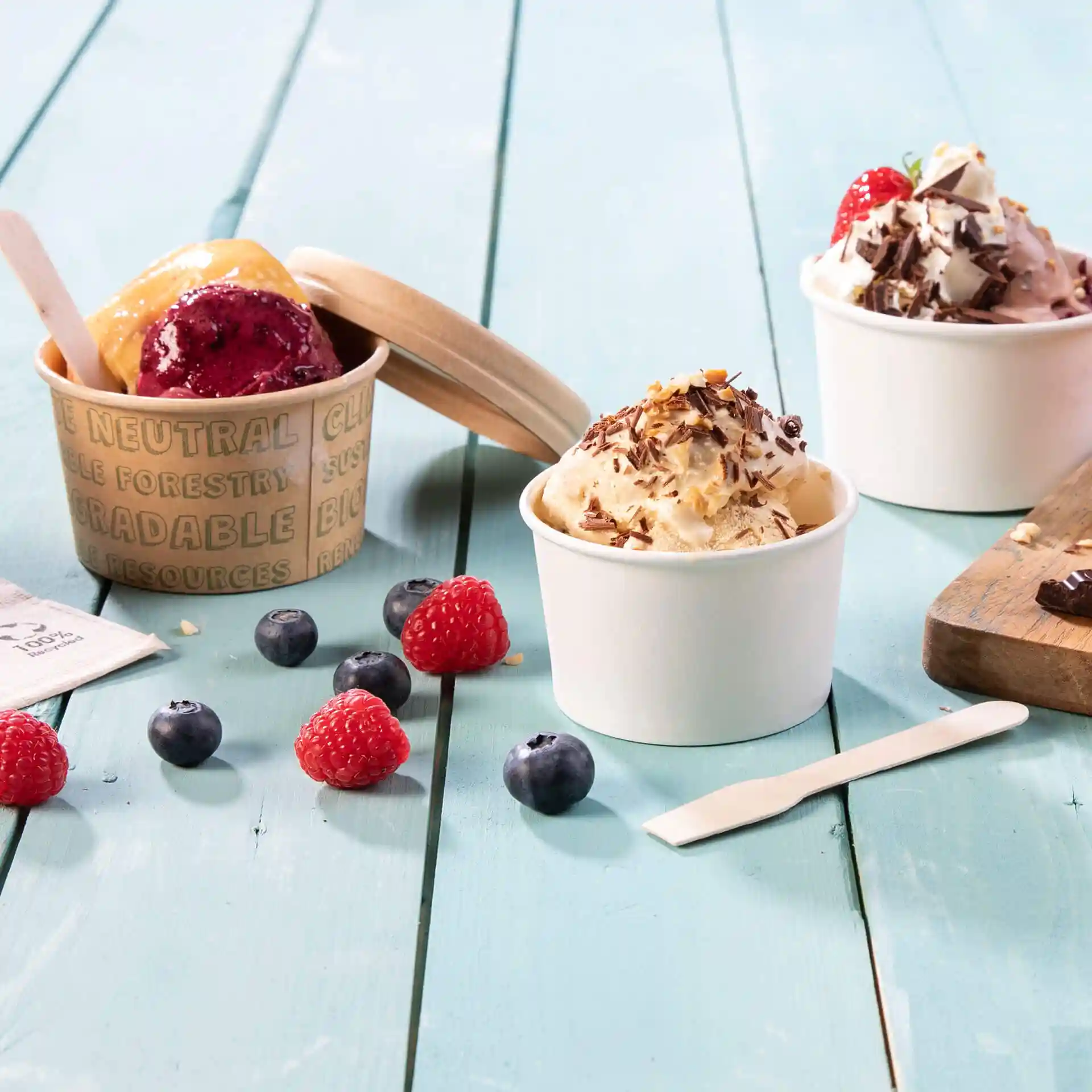 Image of two ice cream cups with different flavors, surrounded by raspberries and blueberries on a blue wooden surface.