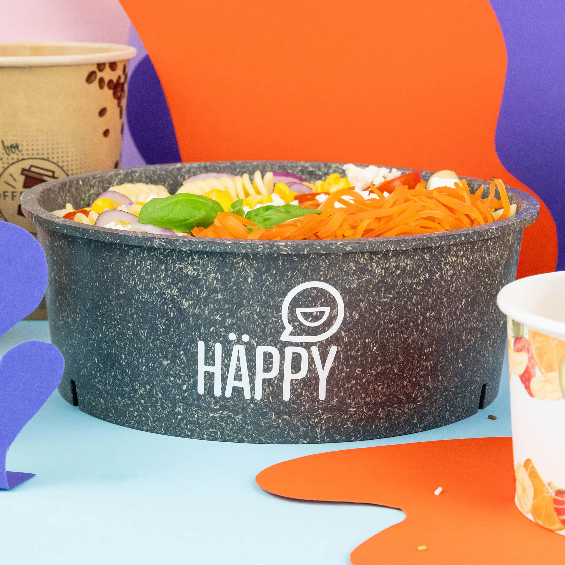 Round reusable bowl in grey with a printed design, filled with pasta and vegetables, on a colorful background.