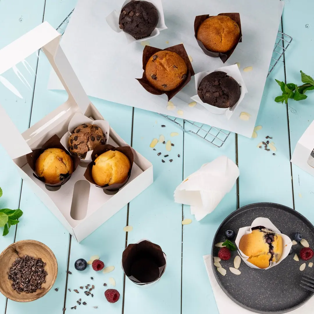 White box containing three muffins, surrounded by additional muffins, berries, and baking ingredients on a blue surface.