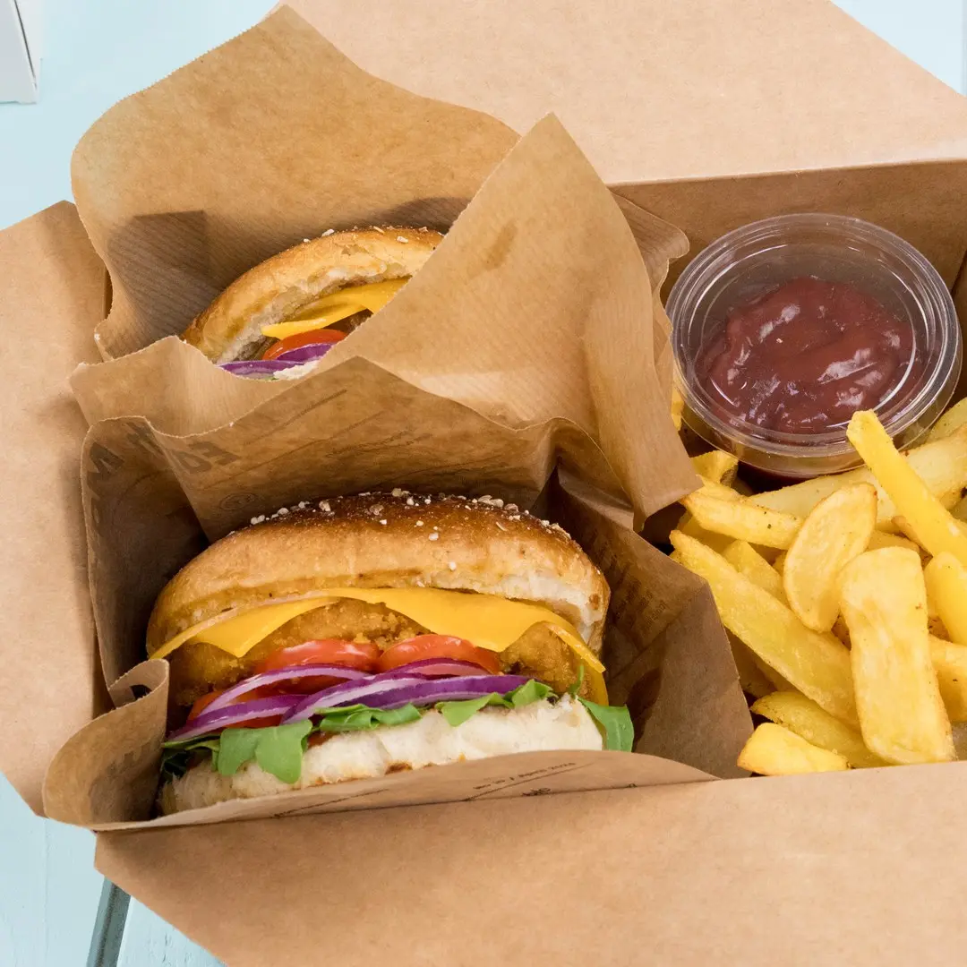 Two burgers with cheese, lettuce, tomato, and onion in a takeaway box, accompanied by fries and a small container of ketchup.
