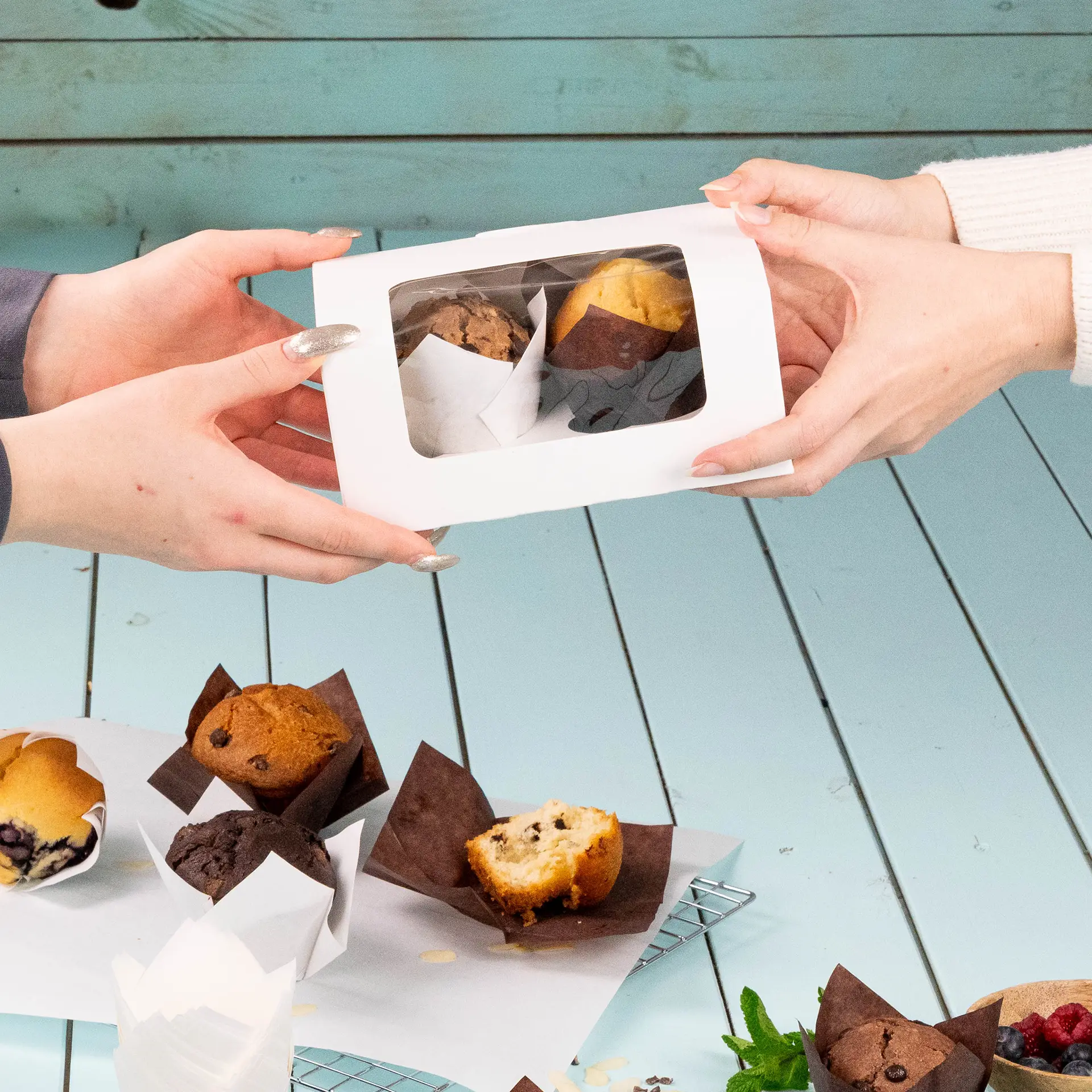 Hands exchanging a transparent box containing two muffins, with several other muffins in brown paper cups on a table.