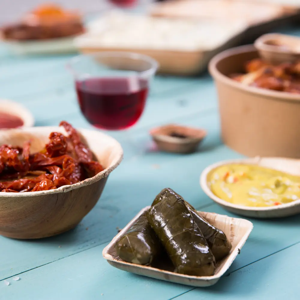 Two small dishes of wrapped food; a bowl of dried tomatoes, a cup of dark beverage, and condiment containers on a blue surface.