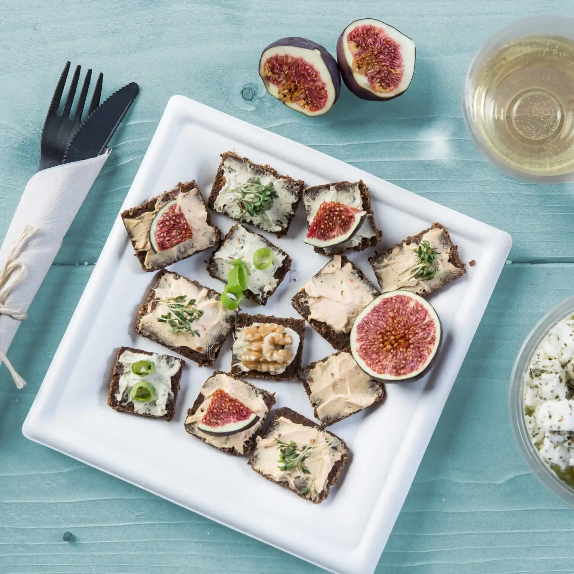 Plate with assorted appetizers including bread topped with spreads, figs, and herbs, on a square white sugarcane plate.