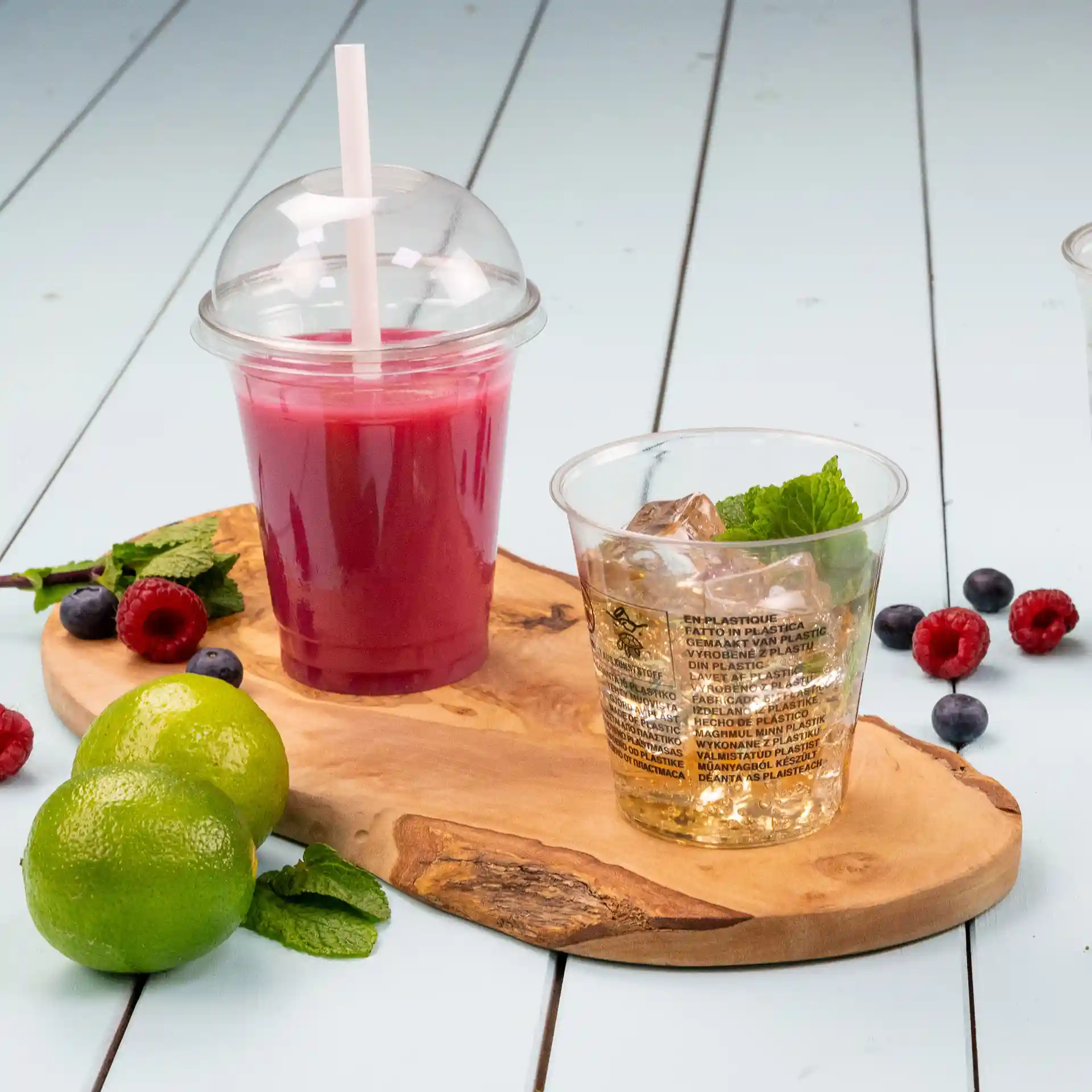 Image of a wooden serving board with a transparent cup of pink drink and a clear cup with ice and mint, surrounded by limes and berries.
