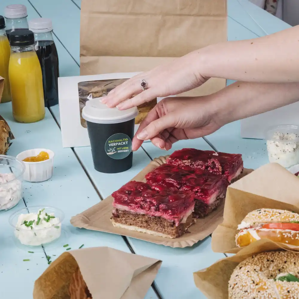 A hand reaching for a small black coffee cup on a table with various food items; layered cake, sauces, and drinks nearby.