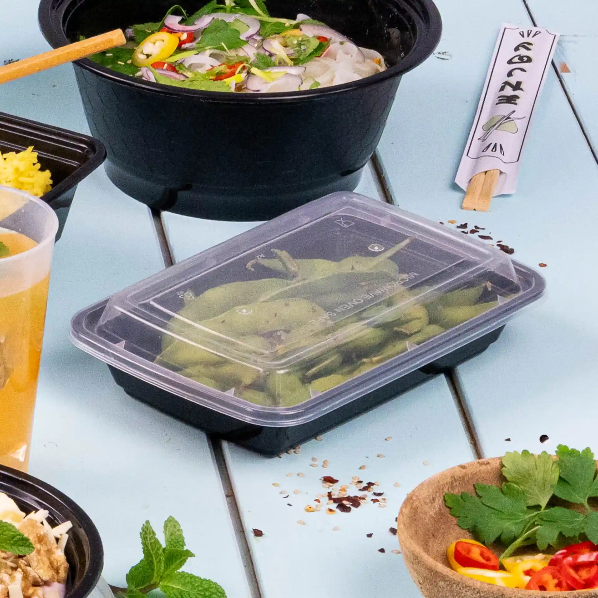Black rectangular reusable container with a transparent lid, containing green beans, on a light blue surface.