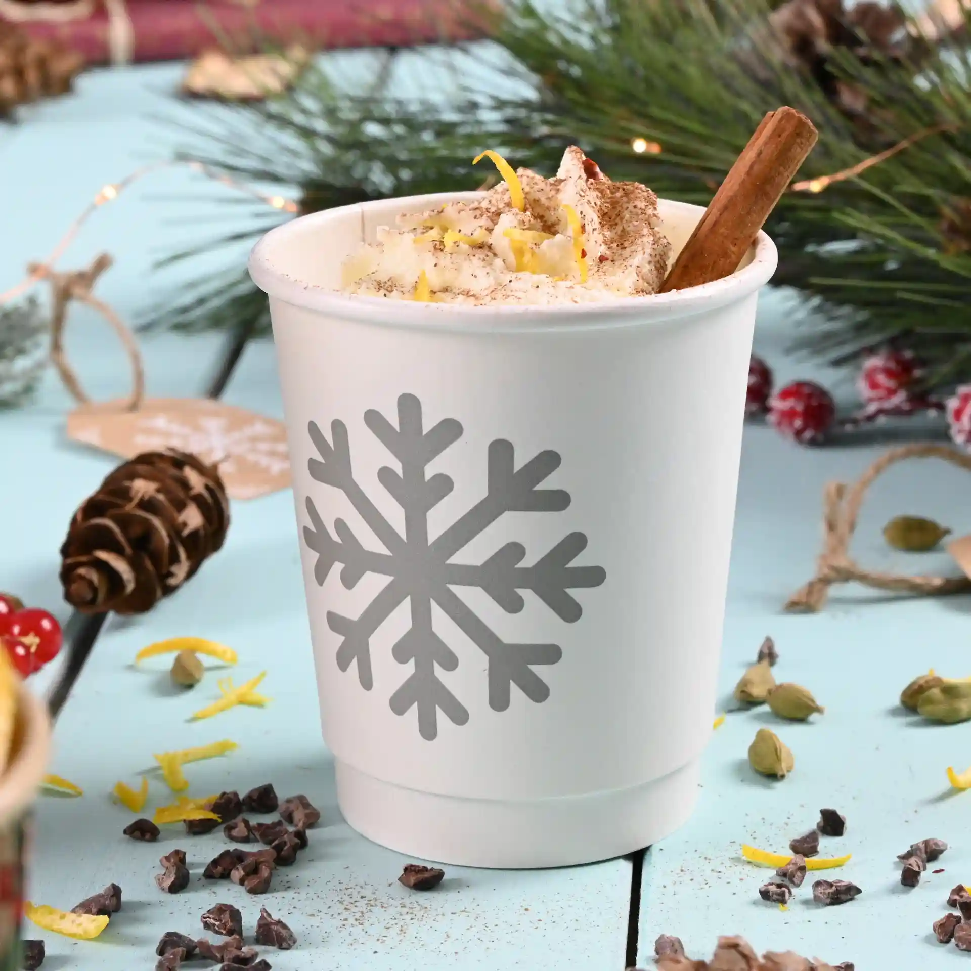White paper cup with a grey snowflake design, filled with whipped cream and a cinnamon stick, on a blue surface.