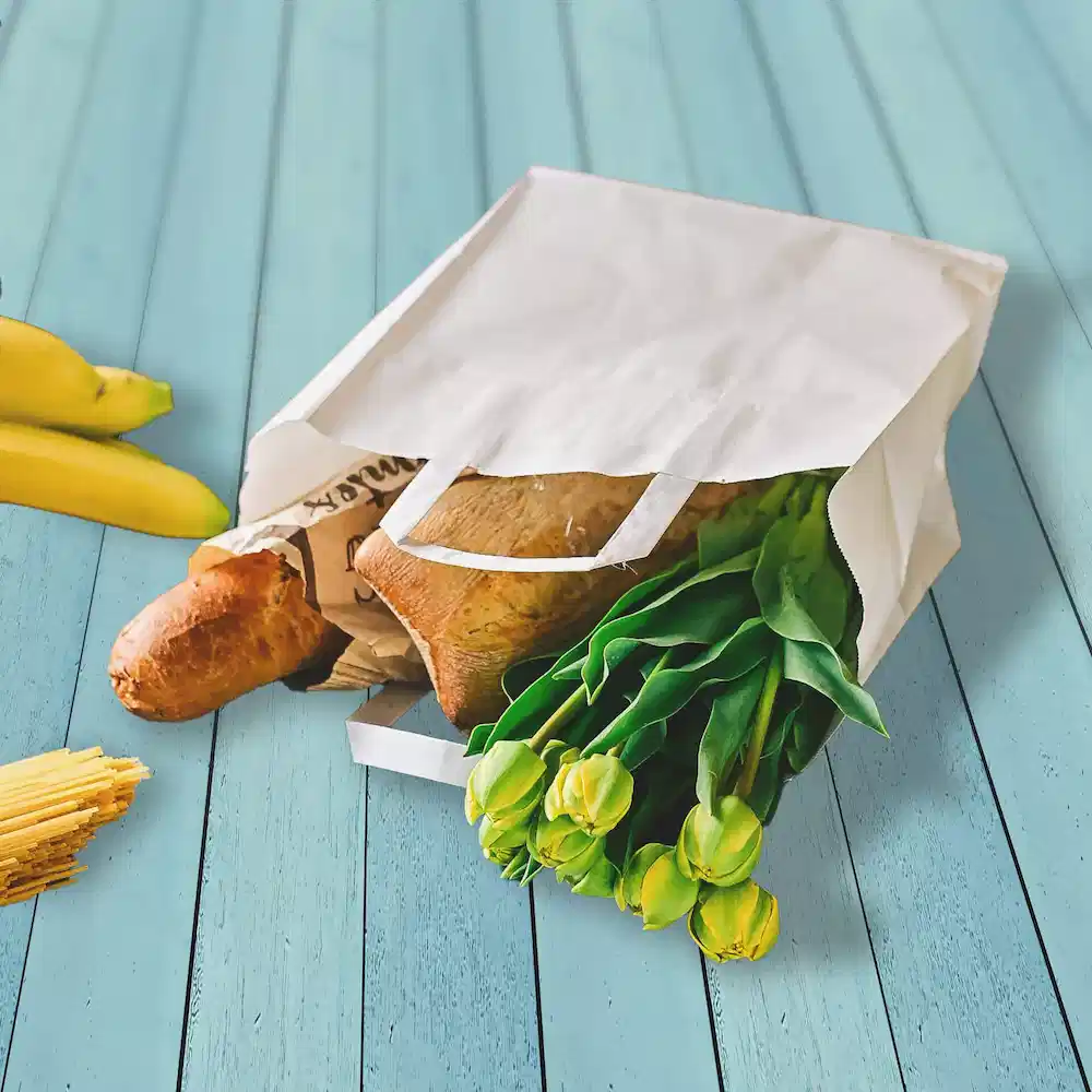 White paper bag filled with groceries including bread, tulips, and bananas on a blue wooden surface.