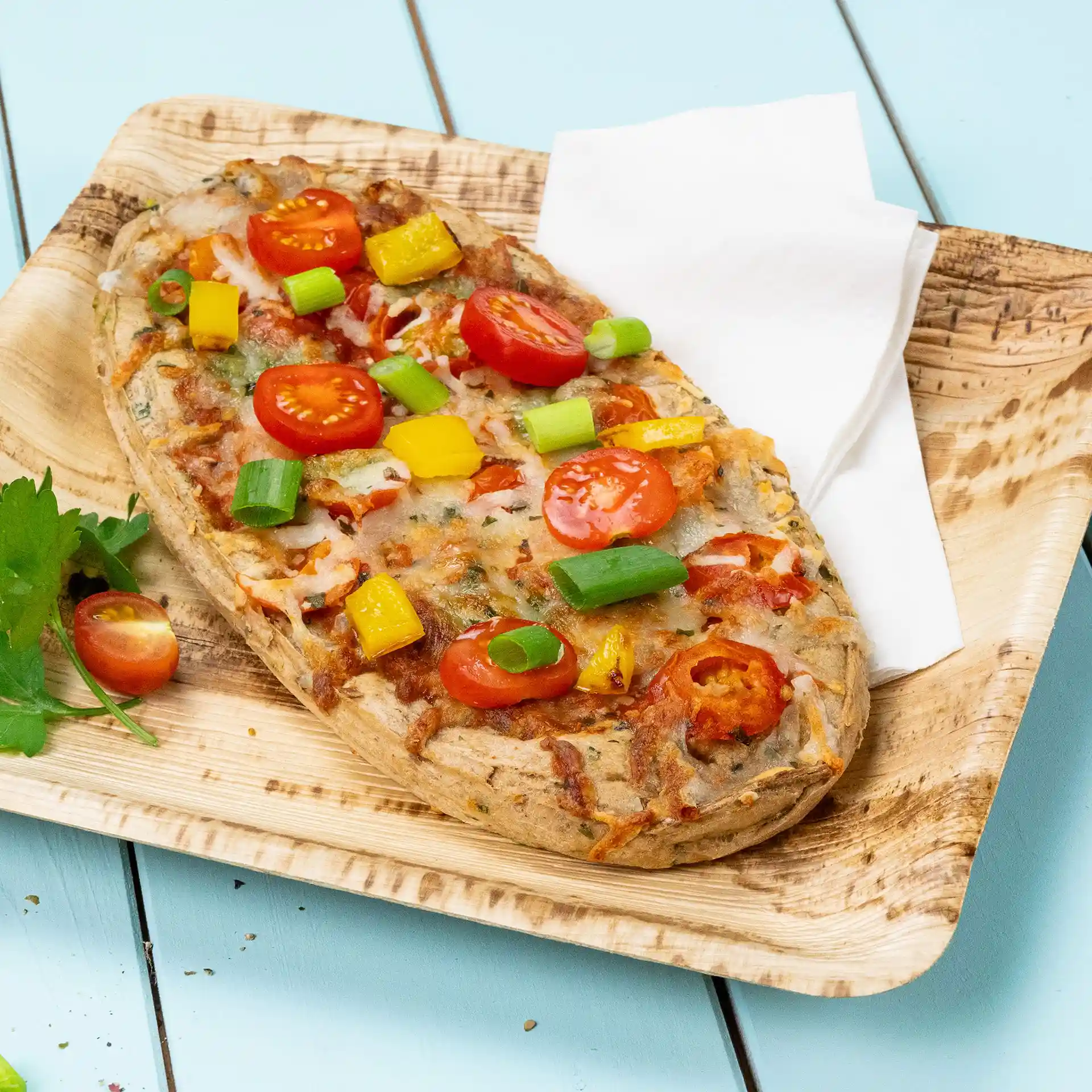 Flatbread topped with cherry tomatoes, yellow bell peppers, and green onions on a wooden plate with a napkin.