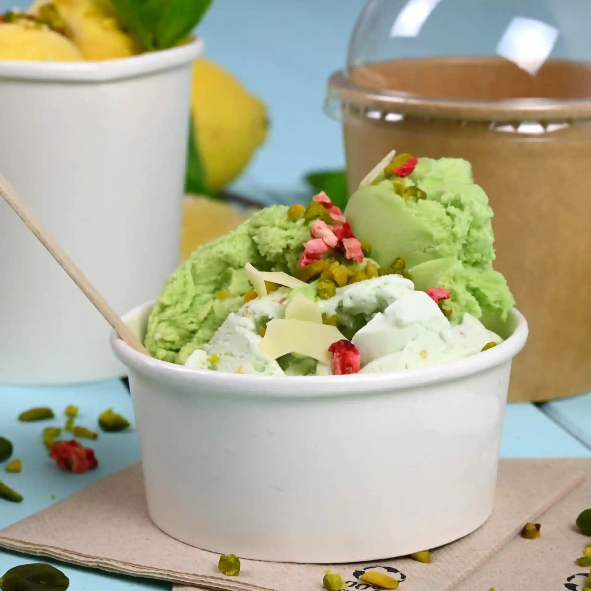 White paper cup filled with green and white ice cream, topped with nuts and pink pieces, on a blue surface.