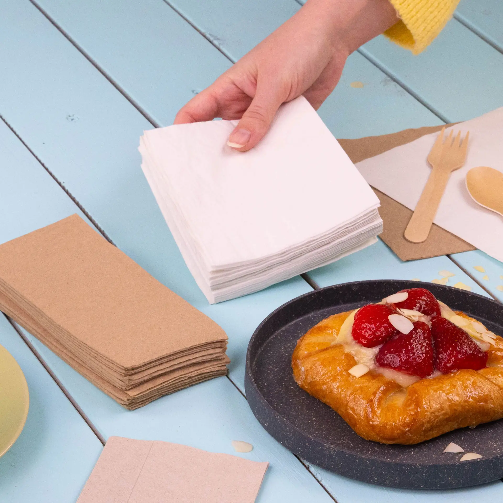 White 2-ply cocktail napkins stacked in a hand, with a croissant topped with strawberries on a plate nearby.