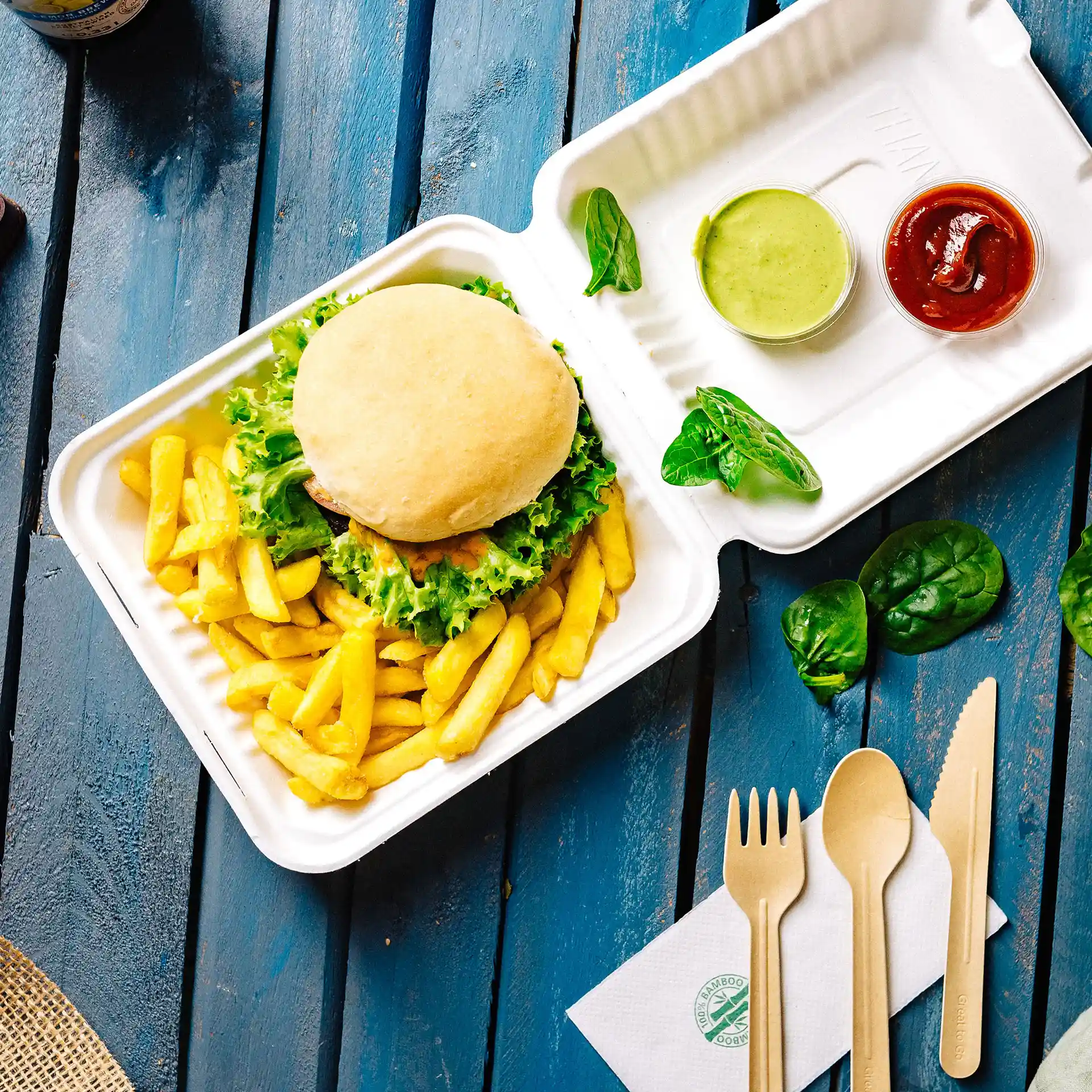 White rectangular hinged-lid container with a burger, fries, green sauce, ketchup, and wooden utensils on a blue surface.
