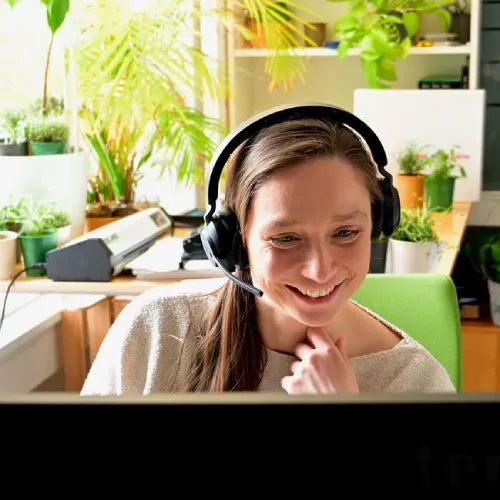Woman with headphones smiling while sitting at a desk with plants and a computer in a bright room.