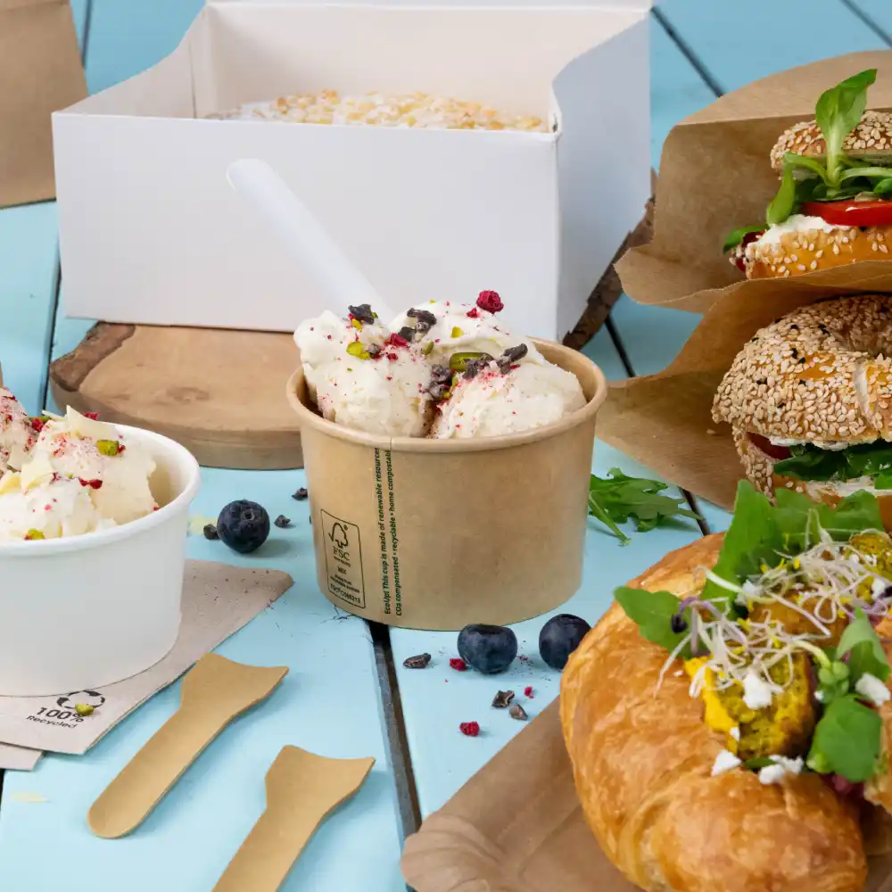 Ice cream in a cup with toppings, two sandwiches on paper, a croissant with filling, and blueberries on a blue wooden surface.