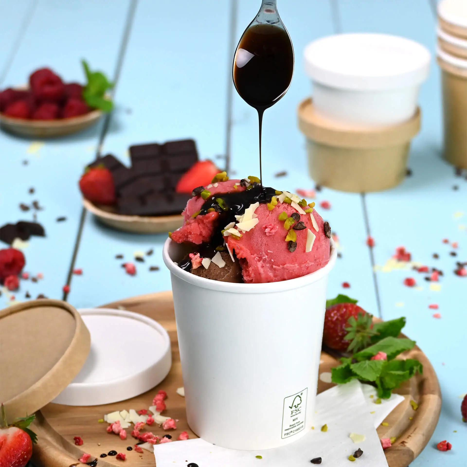 One white paper ice cream cup filled with chocolate and strawberry ice cream, topped with chocolate sauce, pistachios, and shredded chocolate, on a wooden tray surrounded by fresh strawberries, raspberries, and chocolate squares.