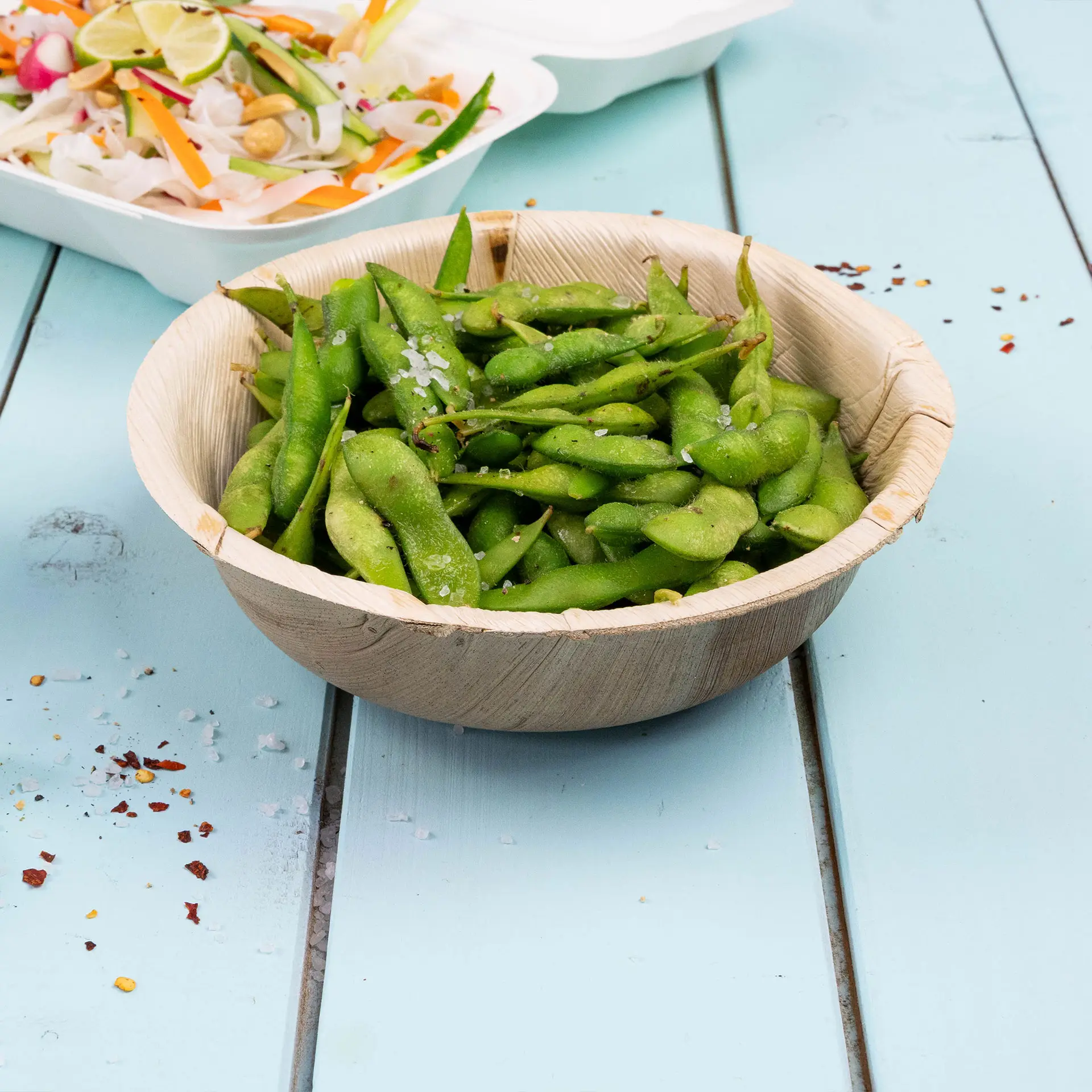 Round palm leaf bowl filled with green edamame, sprinkled with salt, on a blue wooden surface.