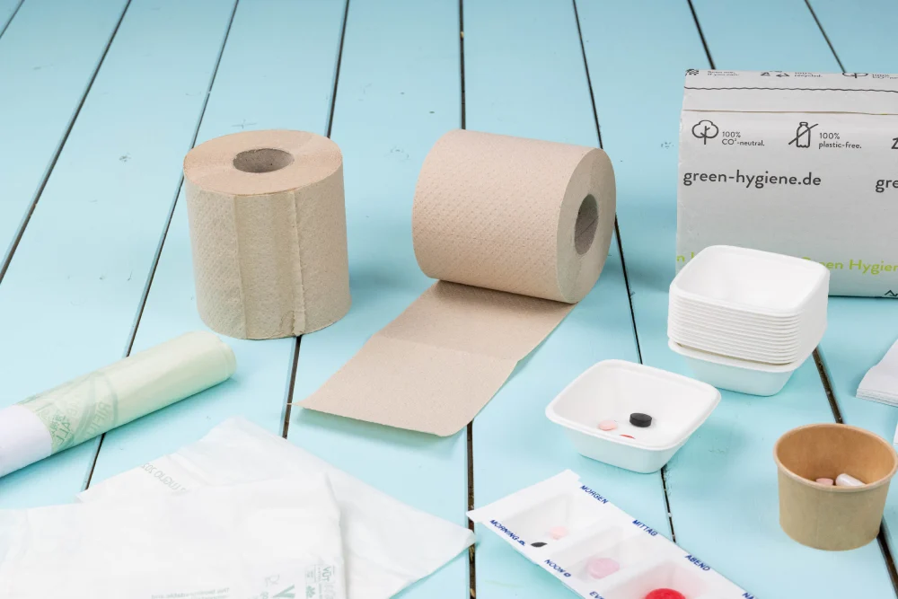Brown paper towel roll and sheet, white disposable containers, compact cardboard box, and assorted tablets on a blue wooden surface.