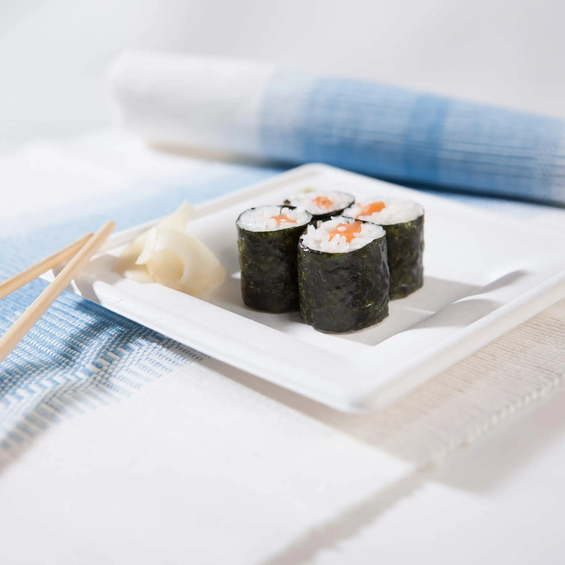 Square sugarcane plate with sushi rolls, pickled ginger, and chopsticks on a textured cloth background.