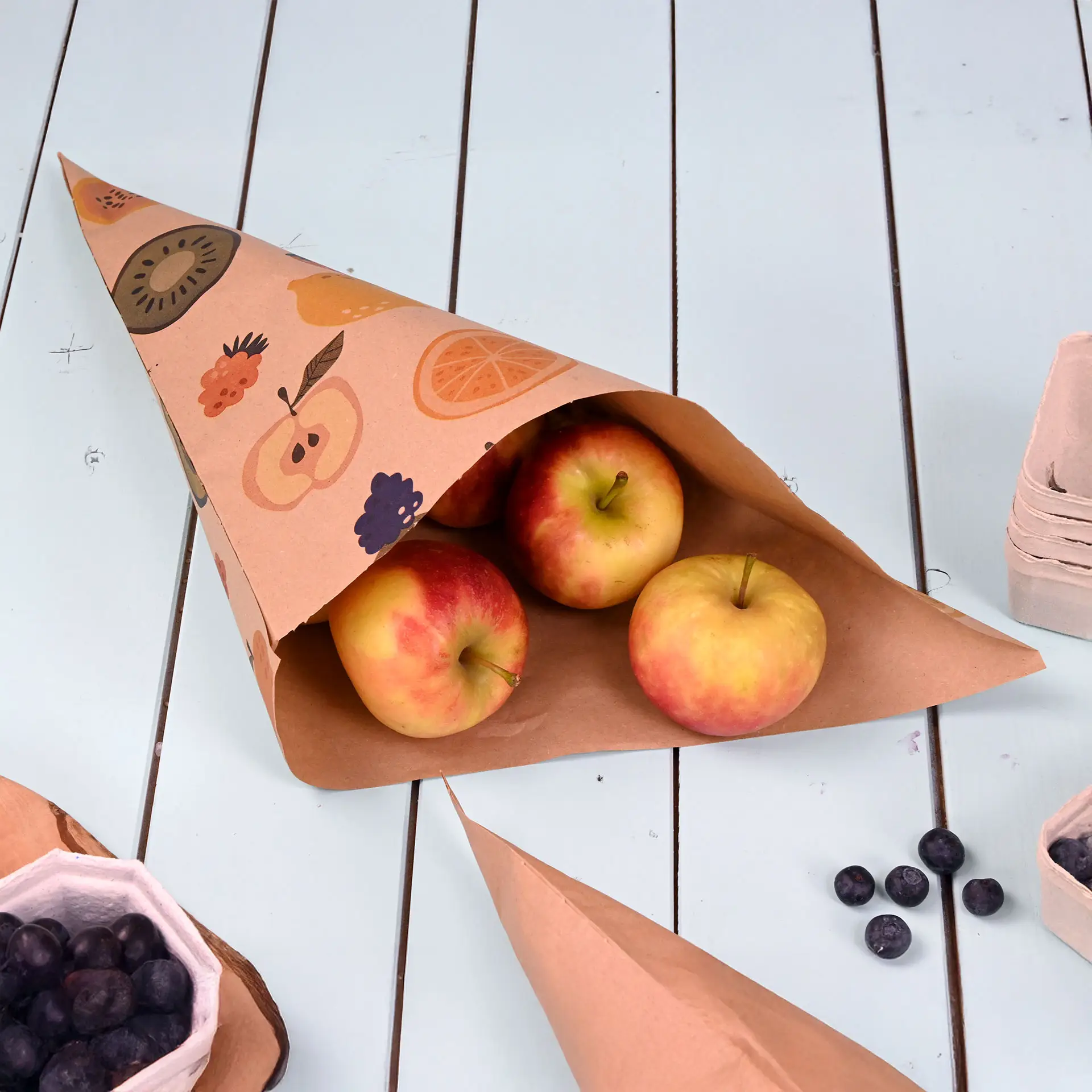 Brown paper cone featuring a fruit design, containing three red apples, on a light blue table surface.