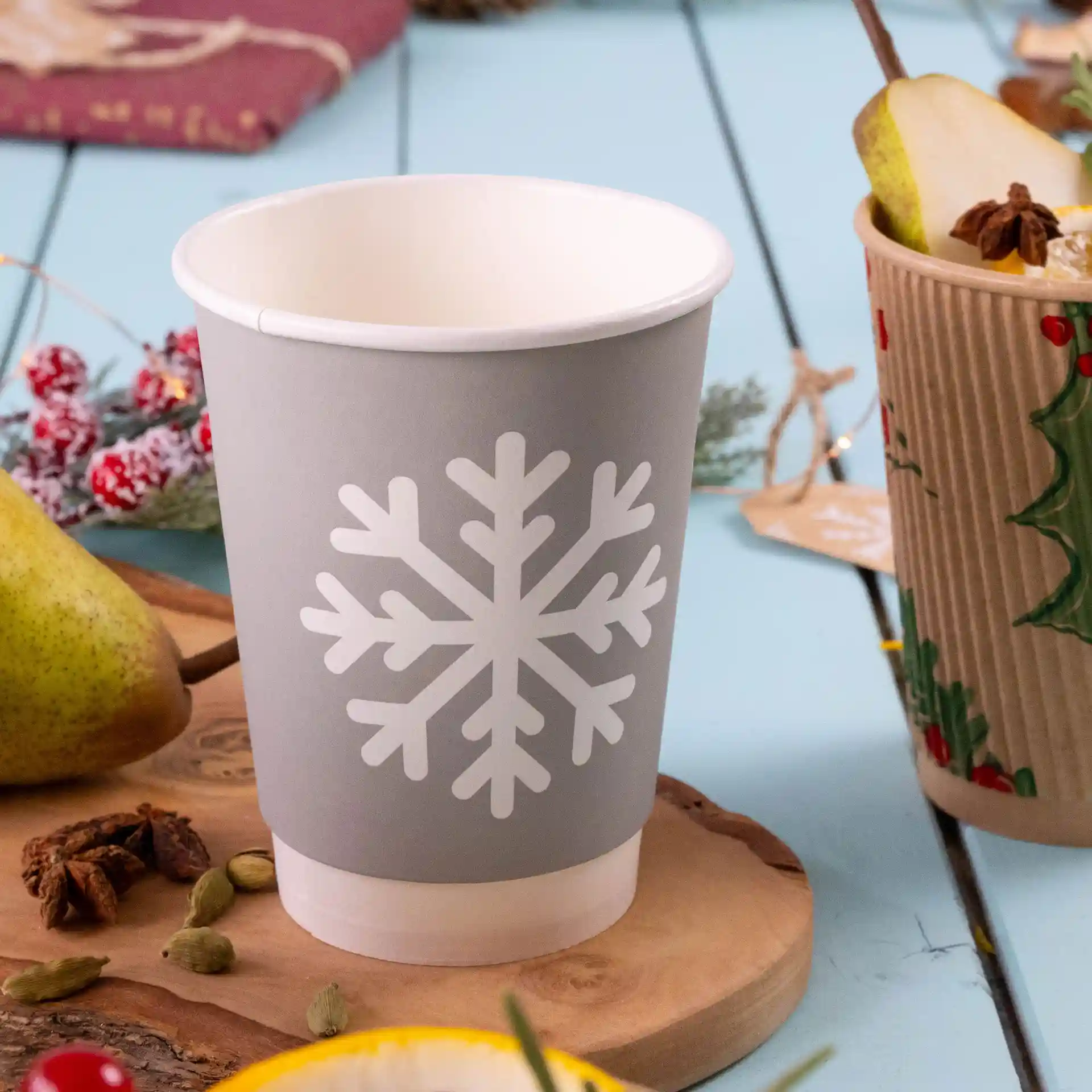 Grey double-walled paper cup with a white snowflake design, 12 oz capacity, on a wooden surface with decorative items.