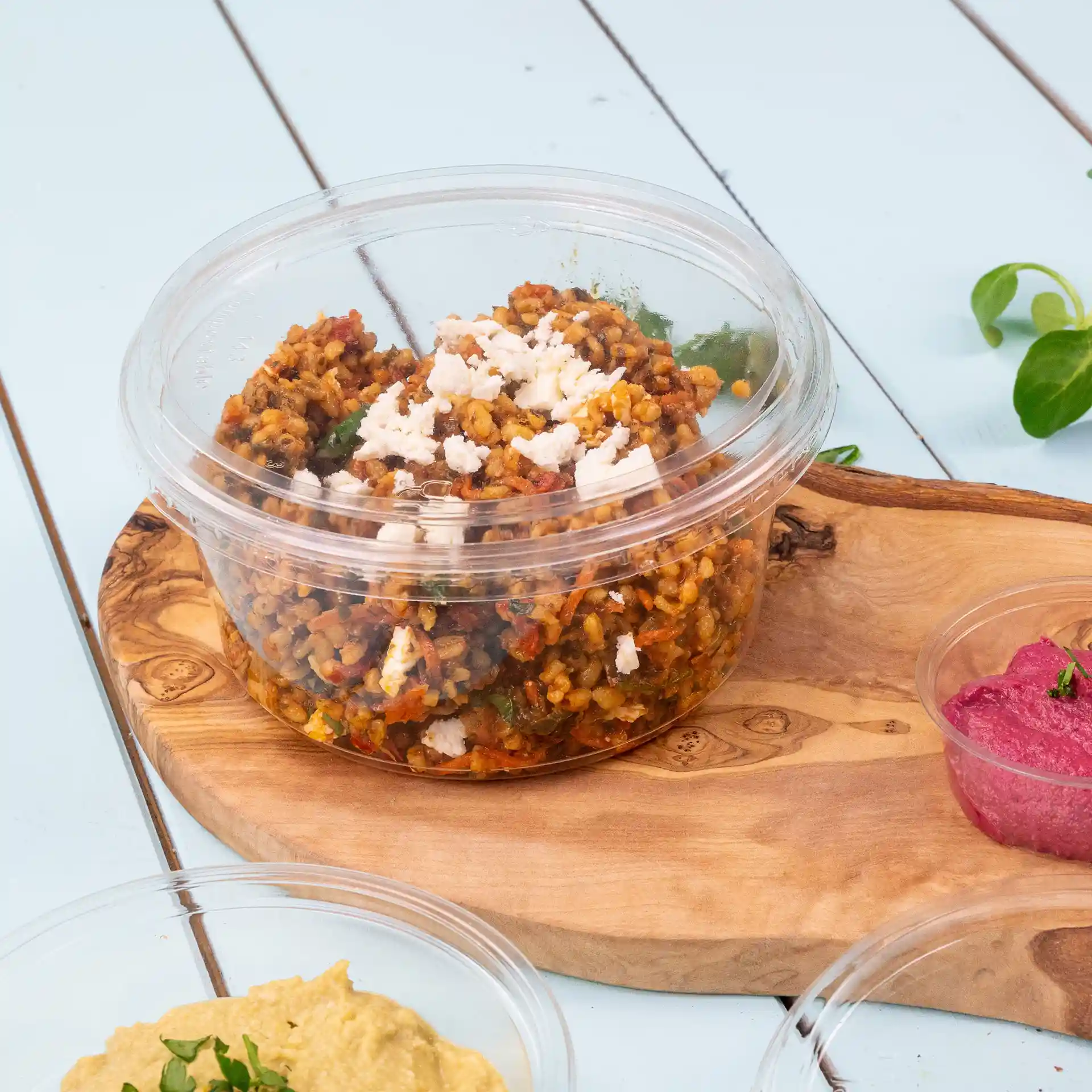Transparent round deli container with a lid, filled with food and placed on a wooden board.