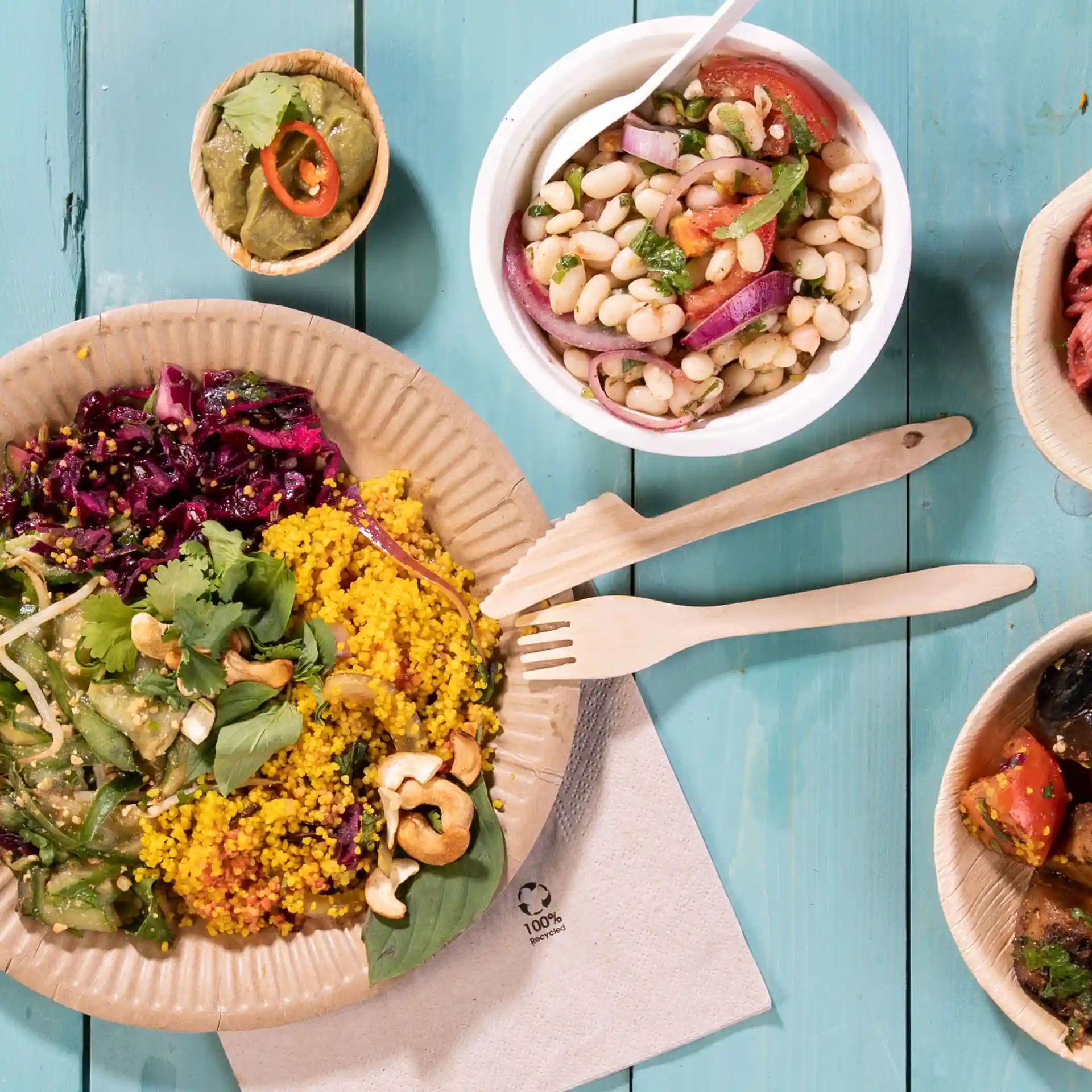 Image of a plate with colorful food including couscous, salad, and a small bowl of beans, accompanied by wooden forks on a blue surface.