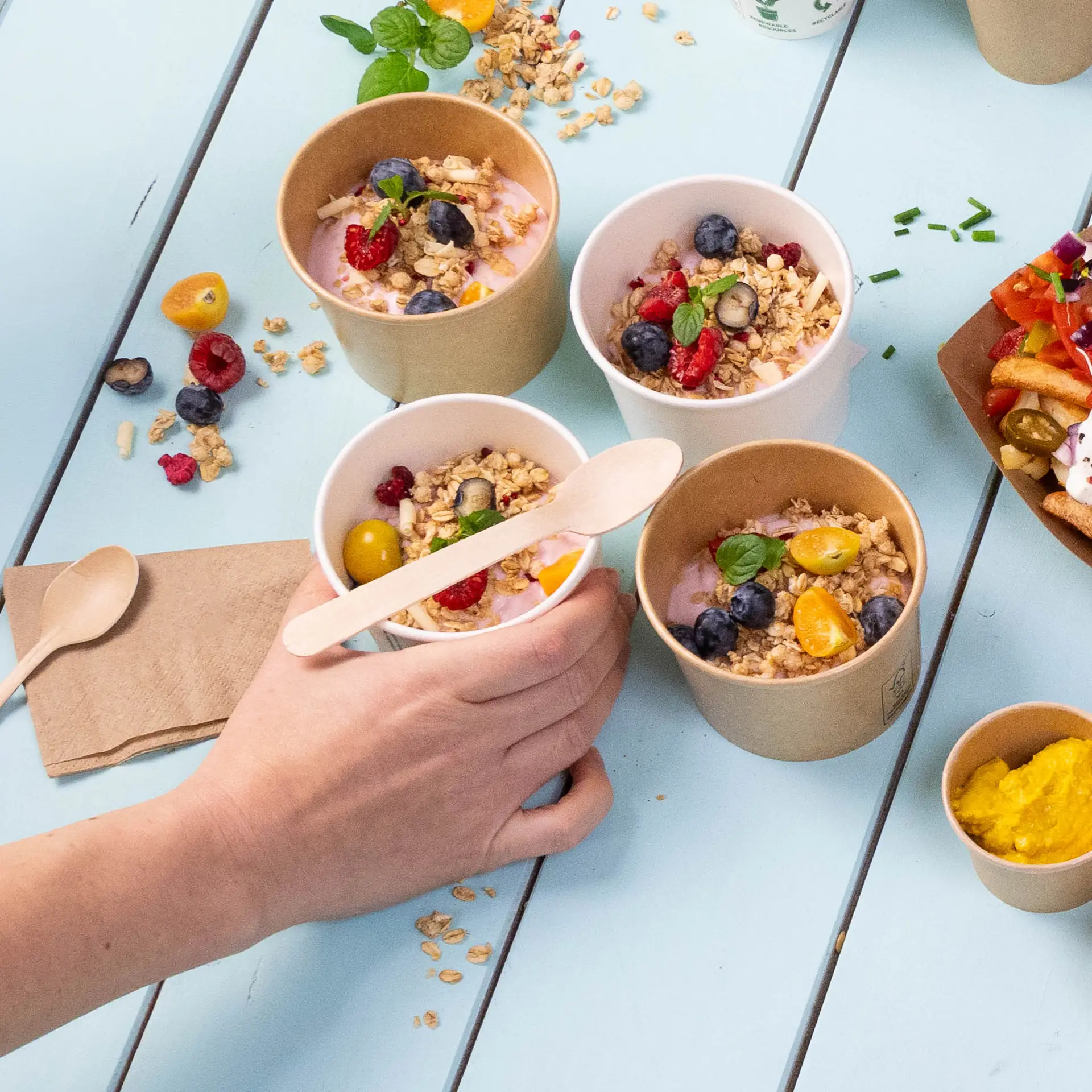 Image of several small cups with yogurt topped with granola and fruits, a hand holding one cup, and wooden spoons nearby.