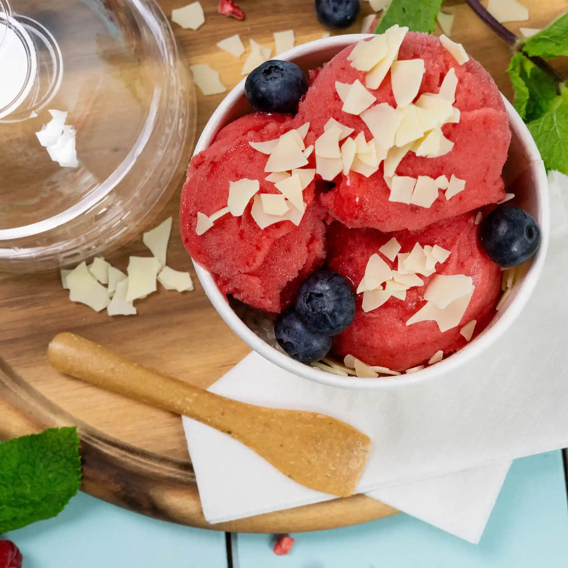 Bowl of red sorbet topped with chocolate shavings and blueberries, accompanied by a wooden spoon on a napkin.