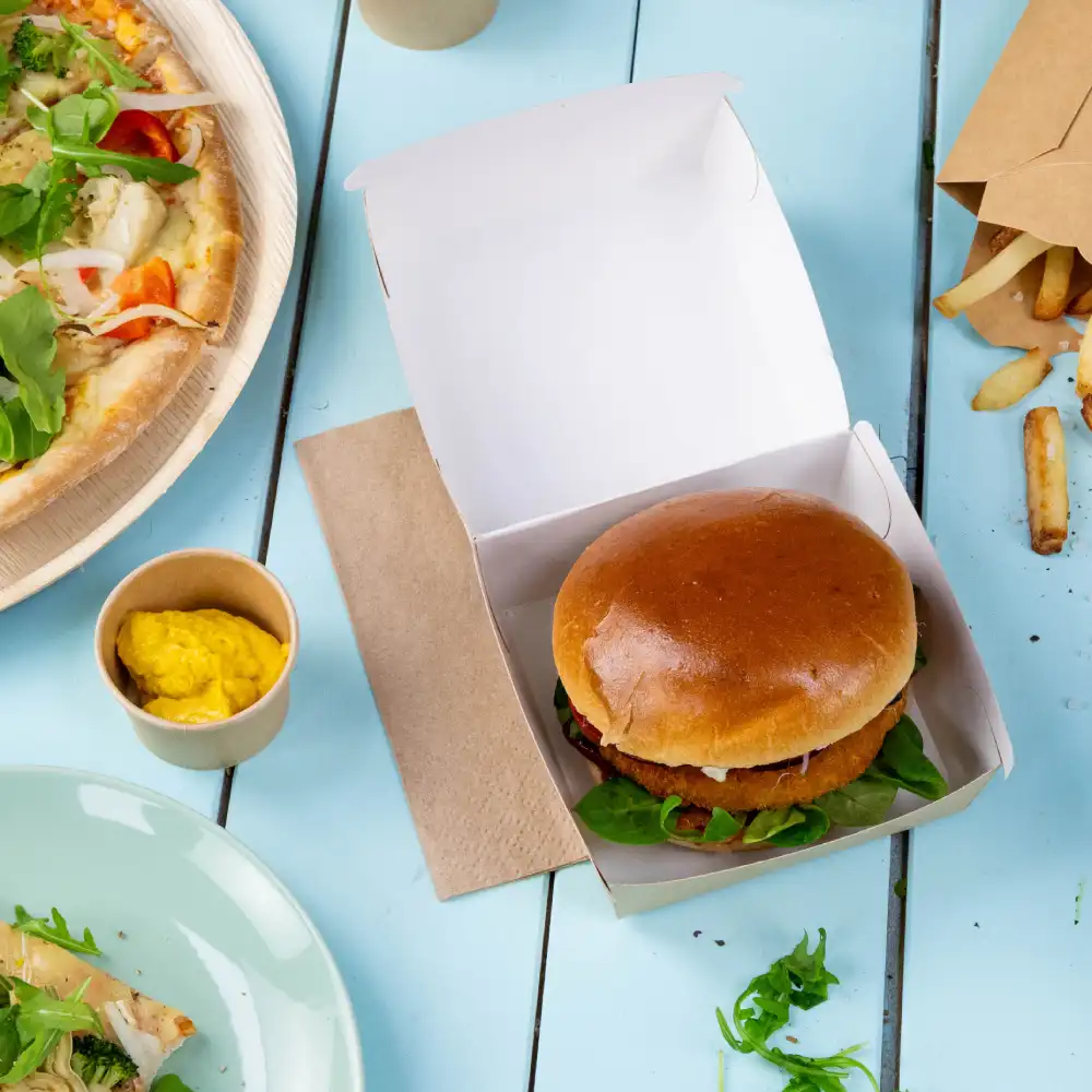 A hamburger with greens is placed inside a takeout box on a blue surface, with pizza, fries, and mustard nearby.