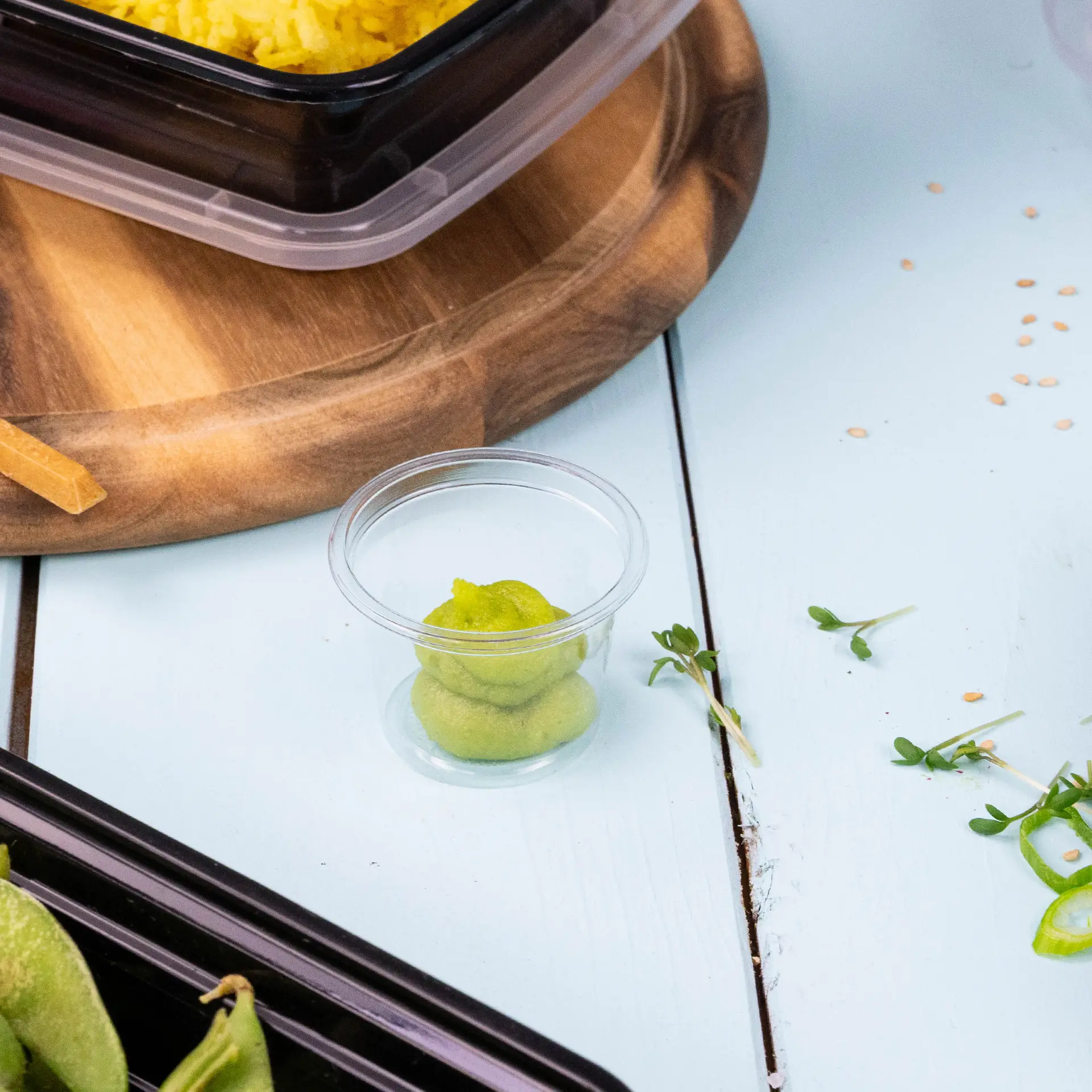 One clear portion cup containing green condiments; surrounded by a black container and salad ingredients on a wooden board.