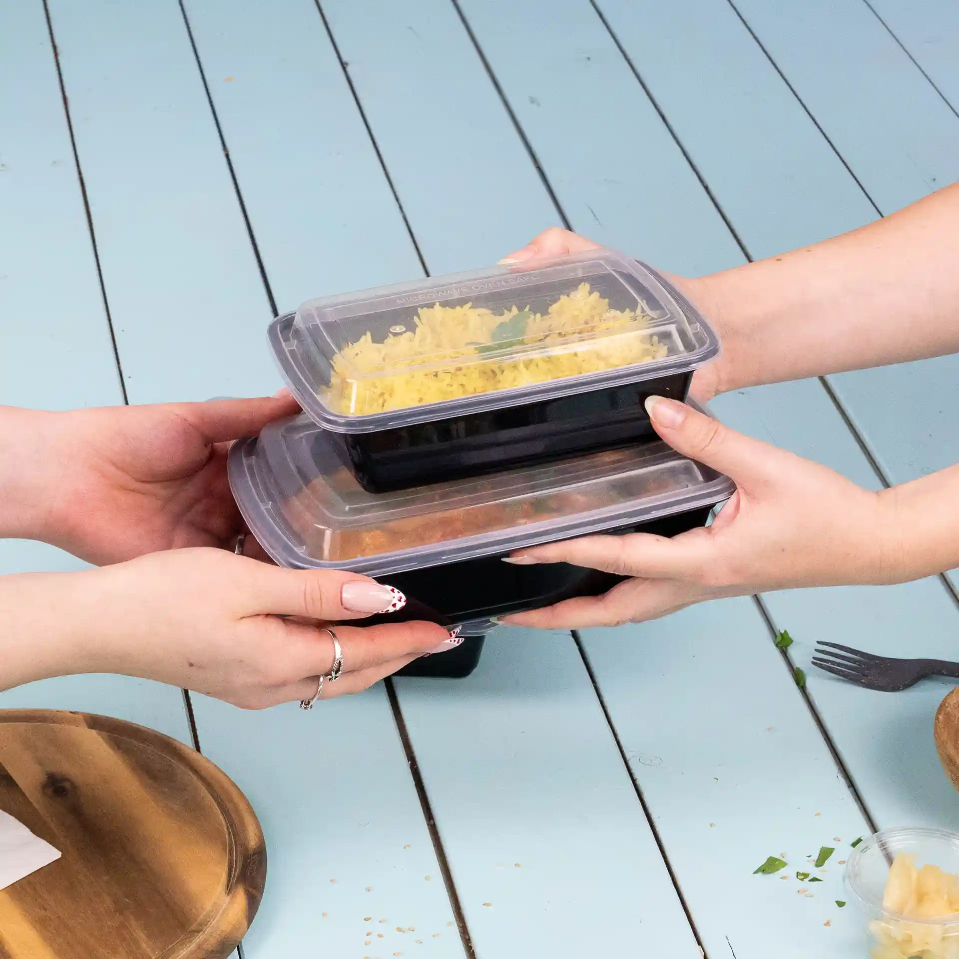 Two hands are holding stacked rectangular food containers with transparent lids, on a light blue surface.