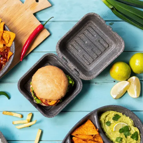 Burger in a reusable kraft box with lettuce and sauces, surrounded by chips, fries, guacamole, and lime wedges.
