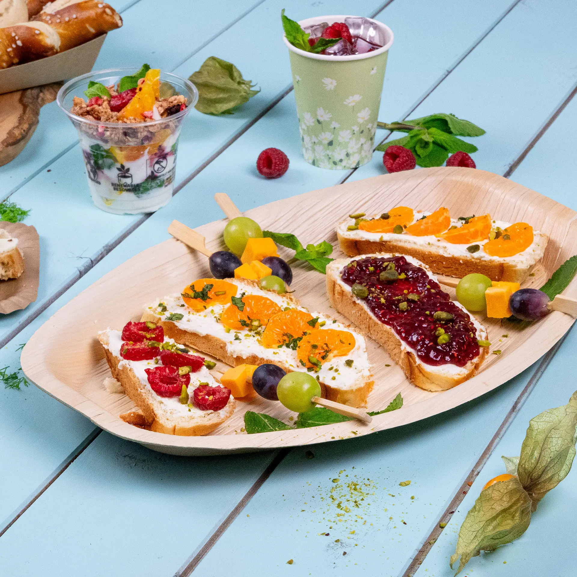 One rectangular palm leaf tray with three slices of bread topped with various spreads and fruit; additional desserts and drinks in the background.