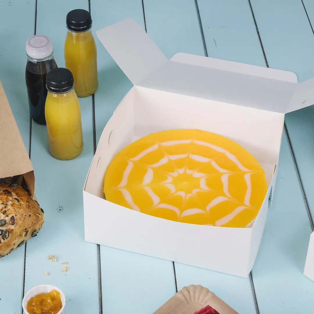 White cake box containing a yellow cake with a white spiderweb design, surrounded by drinks and baked goods.