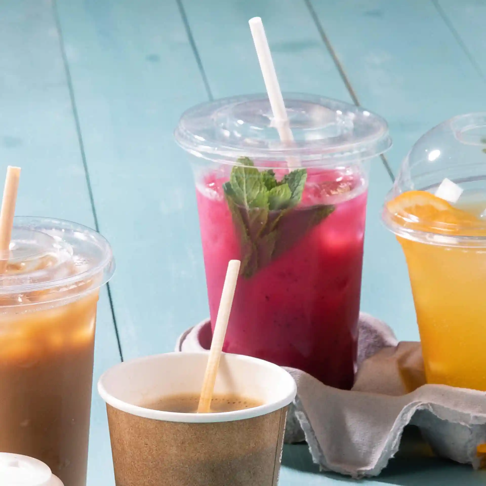 Image of multiple drinks in clear cups with straws, including a pink drink with mint leaves and a brown drink in a paper cup.