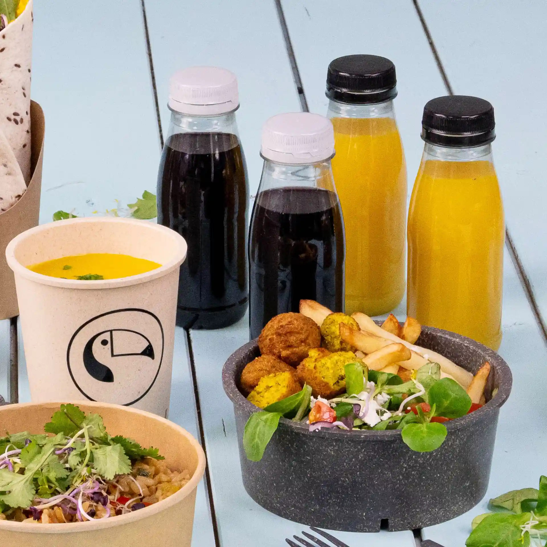 Image of a meal setup featuring a soup cup, two bowls with salad and falafel, and four clear bottles with black and white lids.