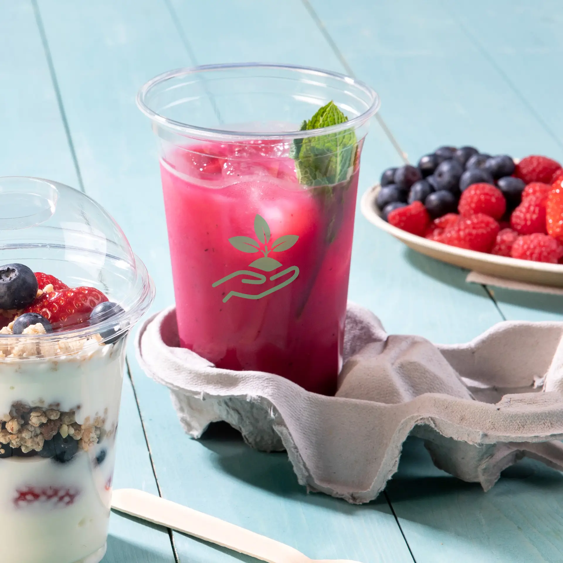 One clear rPET cup filled with a pink drink and a mint leaf; an egg carton holds the cup; a yogurt parfait and berries are nearby.