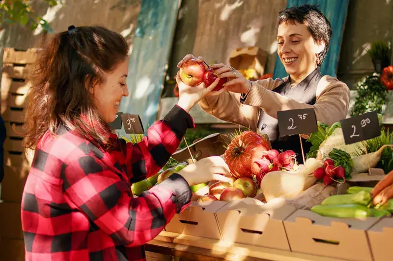 Woman offering apples to a customer at a market stall filled with various fruits and vegetables.