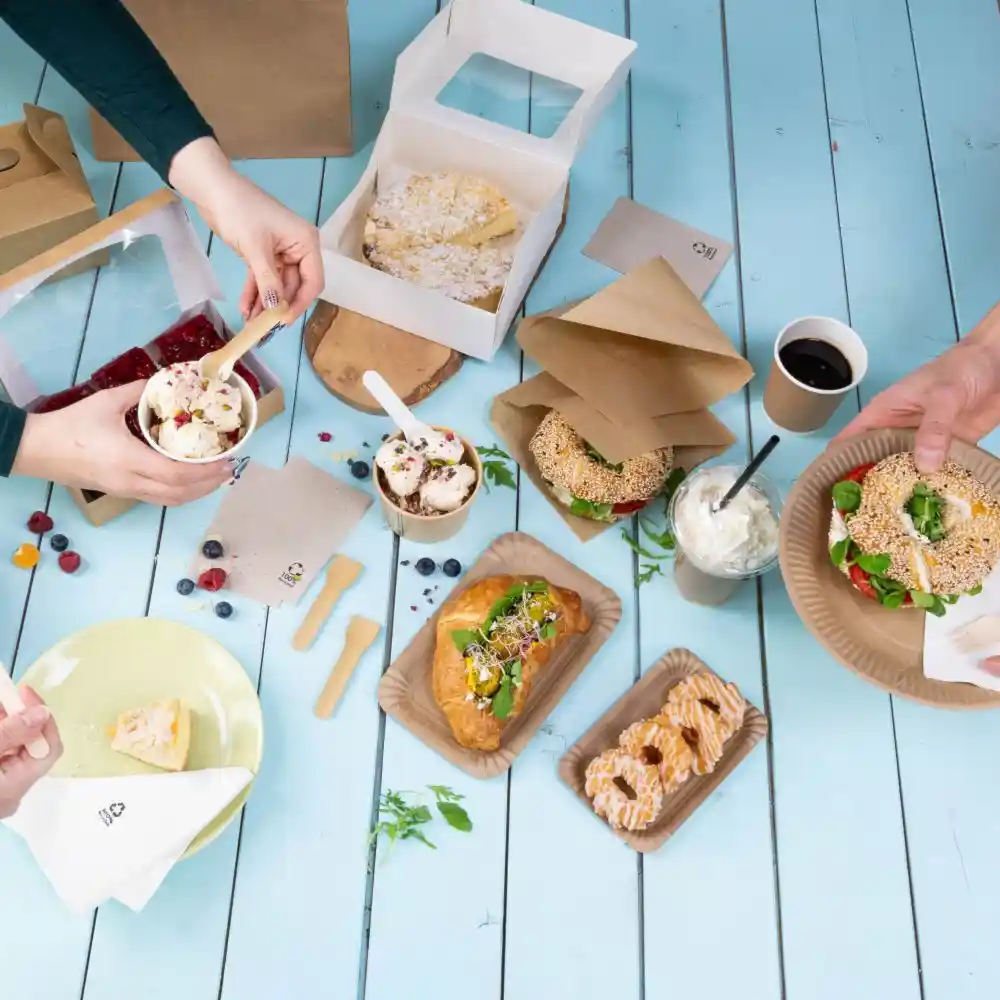 Two cups of ice cream with toppings; a plate with cake; sandwiches and a drink; packaging and utensils on a blue tabletop.