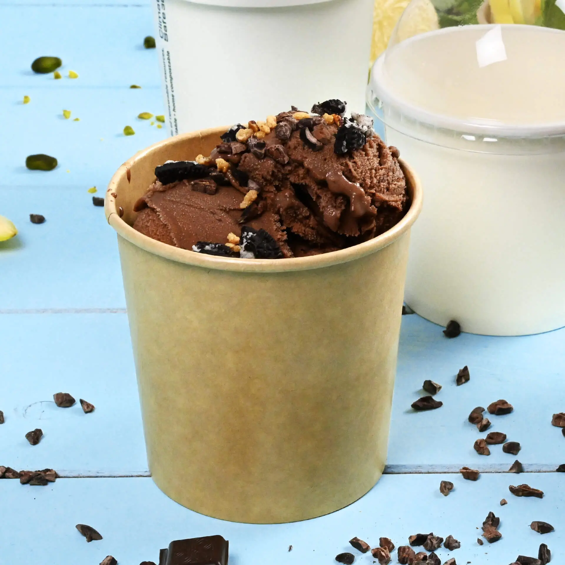 Brown paper ice cream tub filled with chocolate ice cream and toppings, surrounded by chocolate pieces on a blue background.