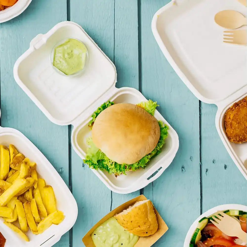 A single hamburger on lettuce in a white takeout container, with fries, dipping sauce, and additional food items visible.