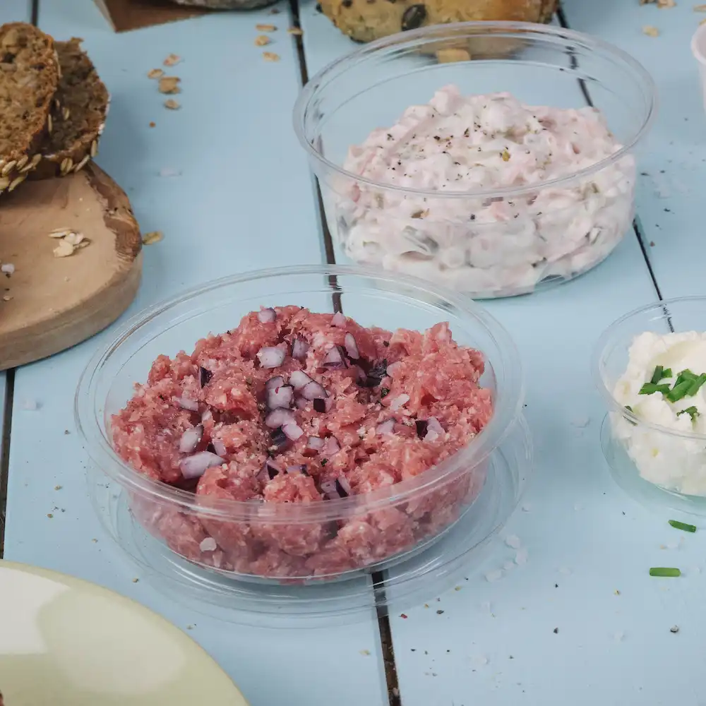 Chopped raw meat with diced onions in a clear container; nearby, creamy dip in another container, and a small bowl of cheese.