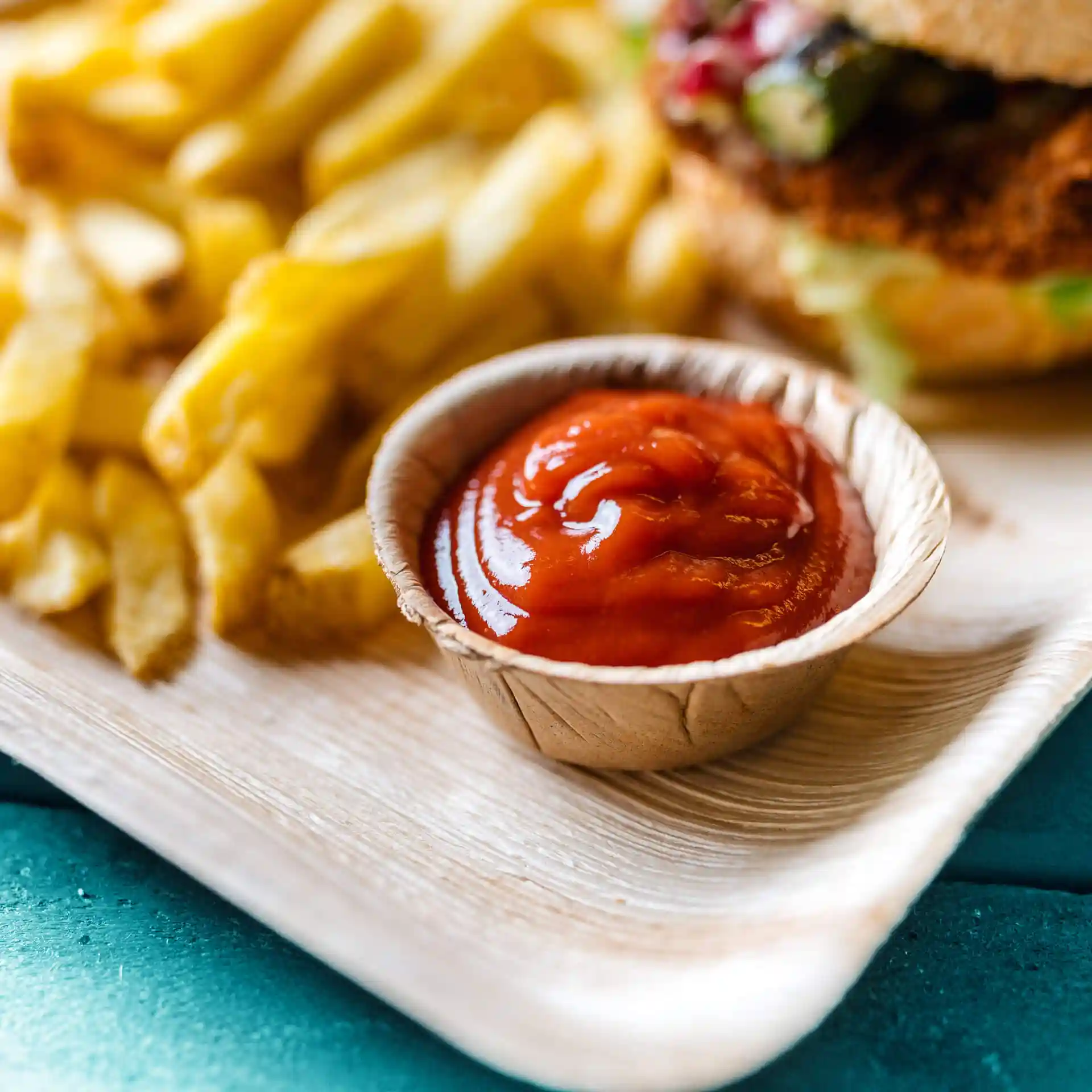 Bowl made of palm leaf containing ketchup, with French fries in the background on a wooden surface.