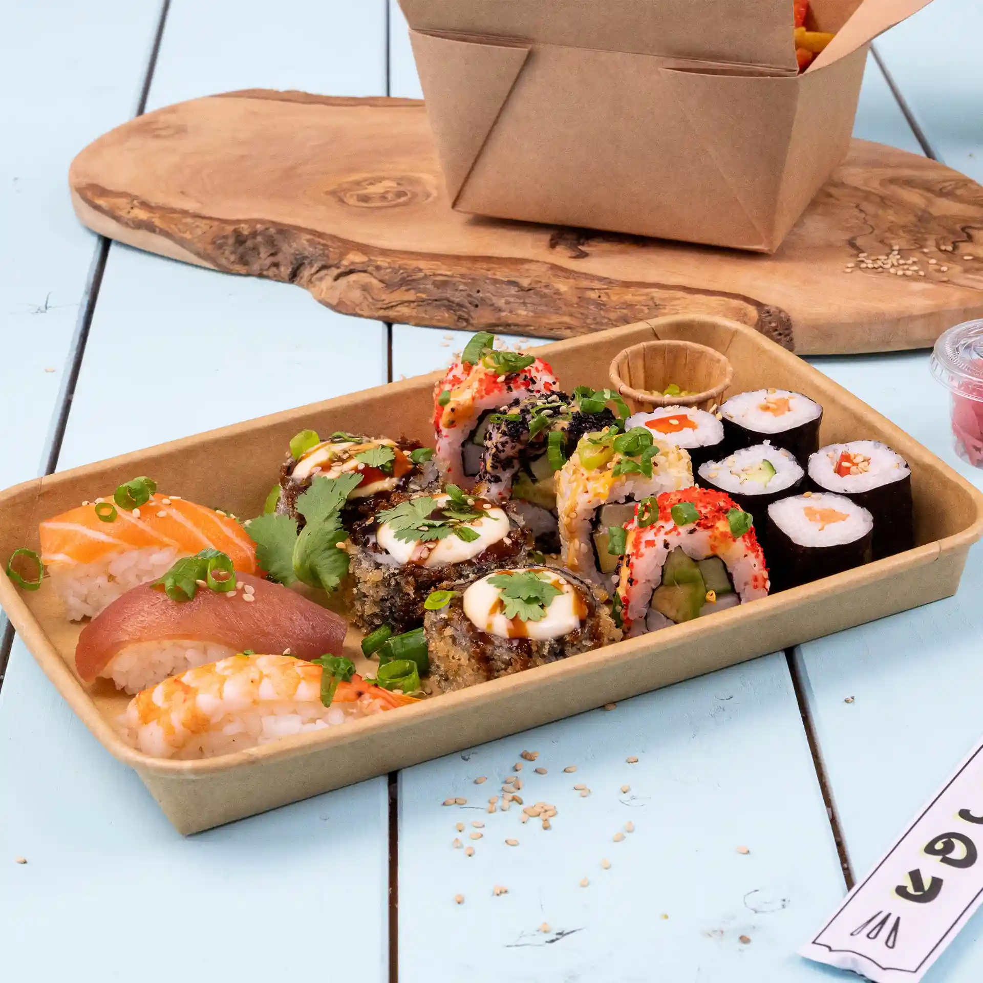 Sushi assortment in a brown PE-coated box, including various rolls and sashimi, on a wooden surface.